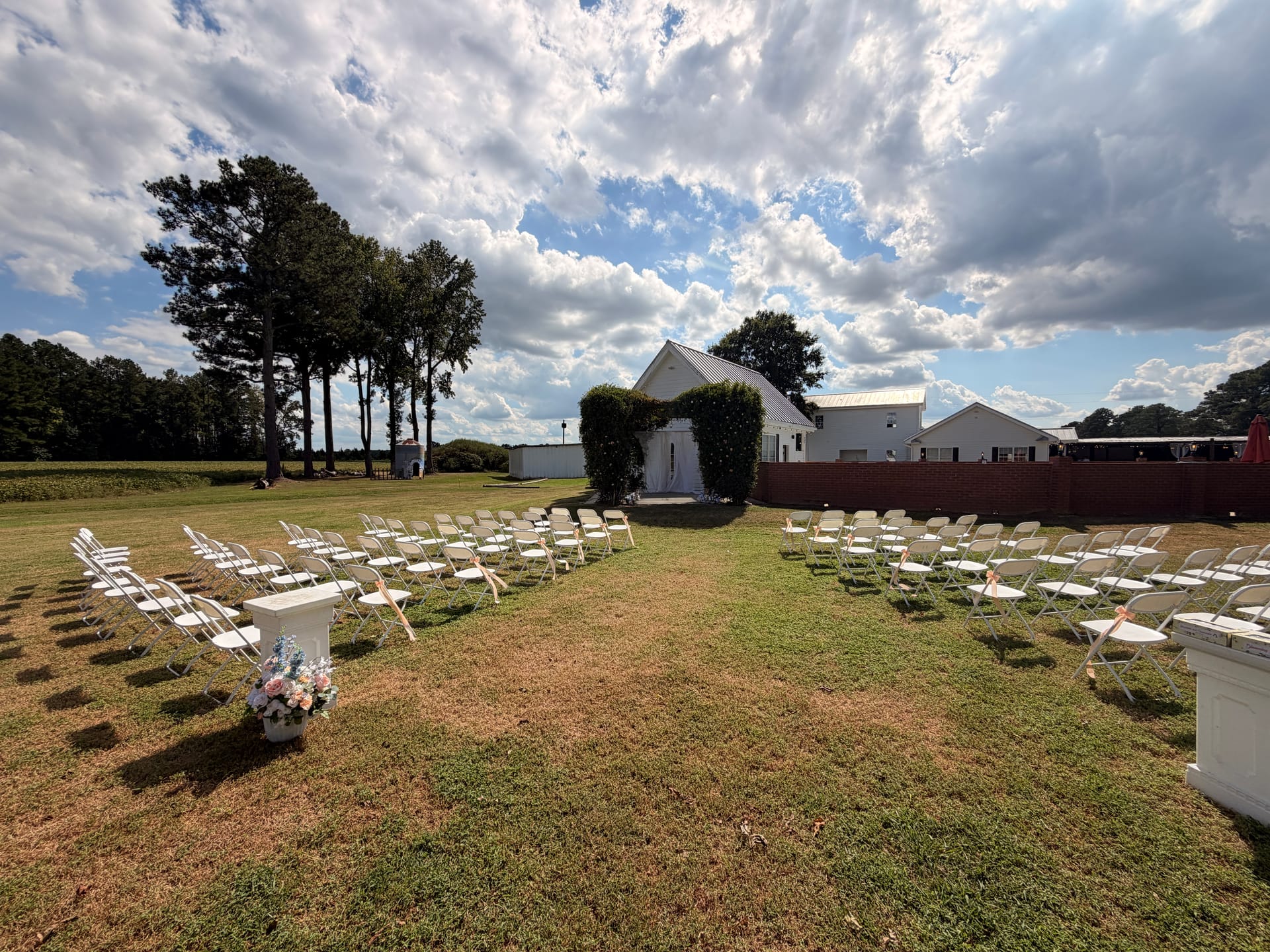 Outdoor wedding ceremony setup at Scarborough House featuring rows of white folding chairs arranged along a grass aisle leading to an ivy-covered chapel under a dramatic partly cloudy sky.