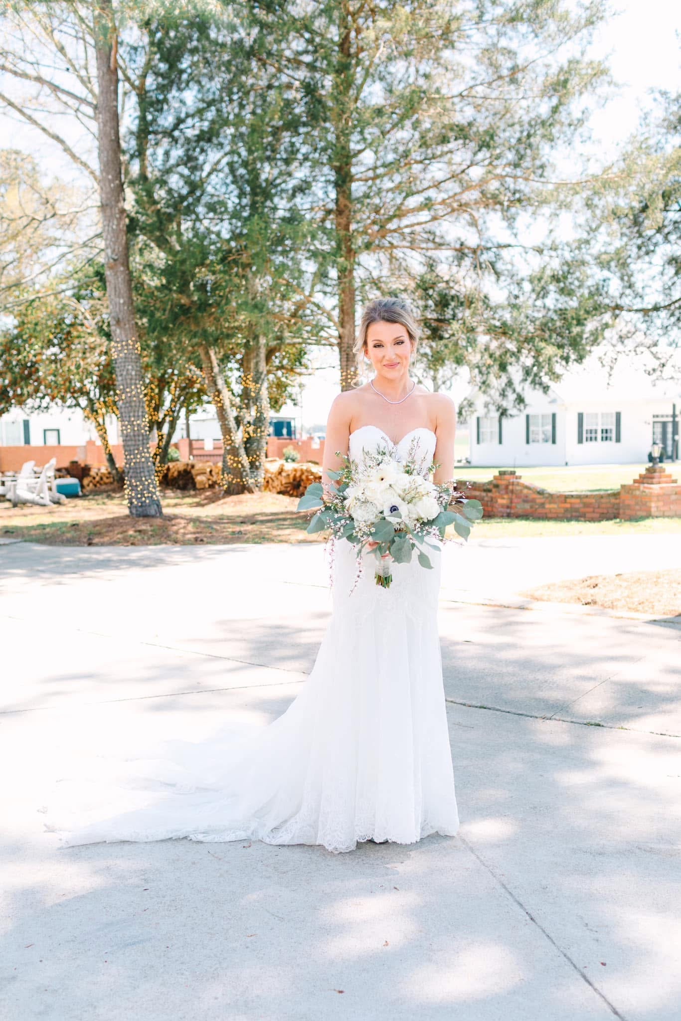 Bride in a strapless white lace gown holding a white and greenery bouquet stands on the driveway of the historic Scarborough House estate surrounded by tall trees and string lights.