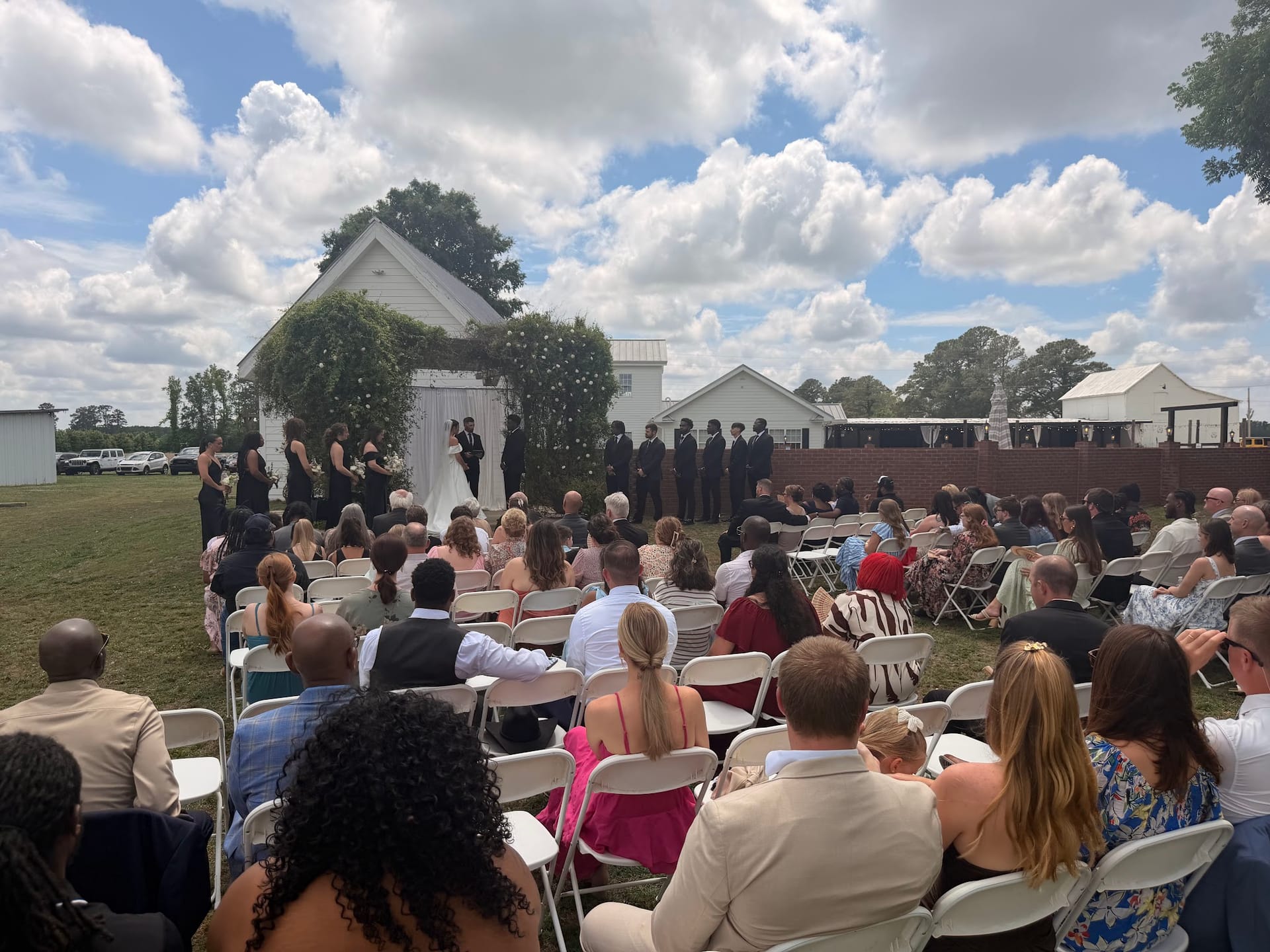 Guests seated in white folding chairs watch a wedding ceremony on the lawn in front of a white chapel at Scarborough House historic estate venue.