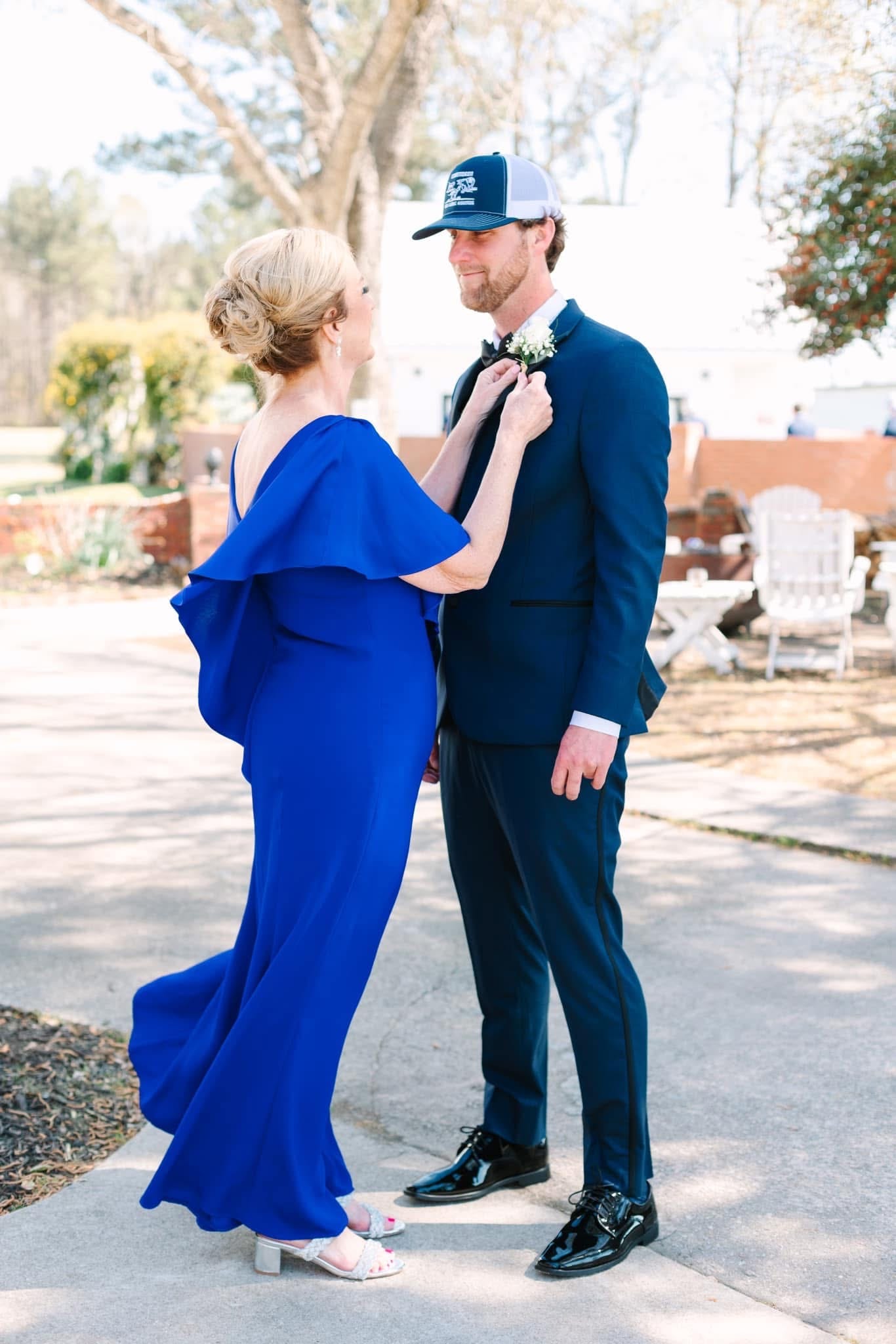 A woman in a vibrant royal blue gown adjusts the boutonniere on a groom in a navy suit and trucker cap on the grounds of a historic wedding venue.