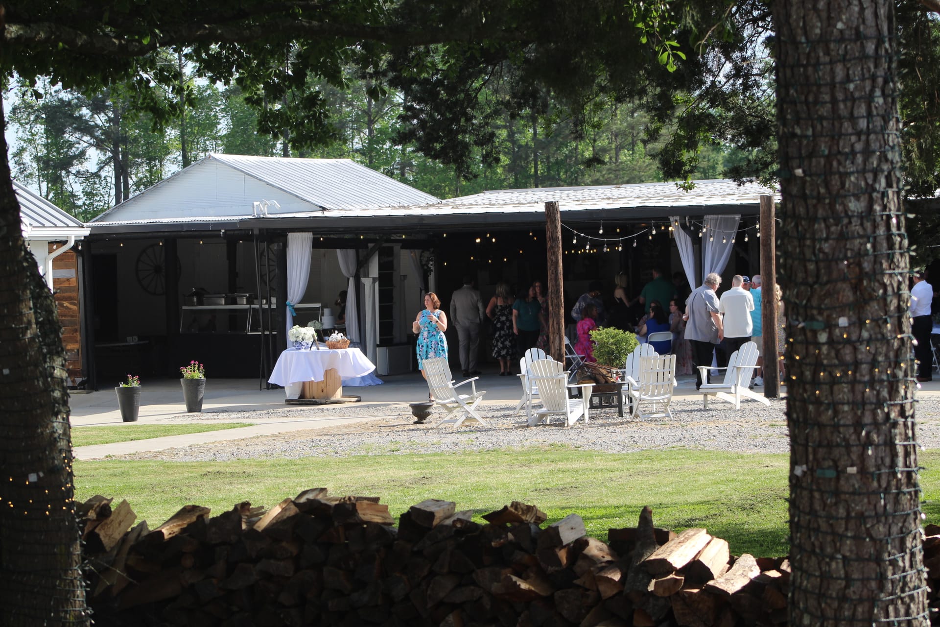 Guests mingle under a string-lit open-air pavilion with white draping and Adirondack chairs arranged on the gravel patio at a rustic wedding reception surrounded by tall trees.