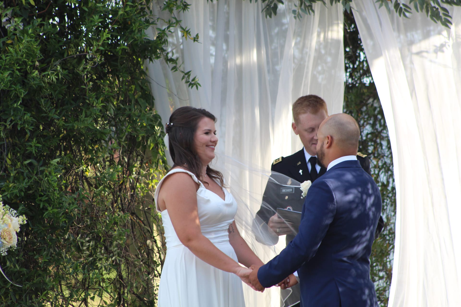 Bride in a white gown smiles joyfully while exchanging vows with her groom under a draped floral arch at an outdoor wedding ceremony.