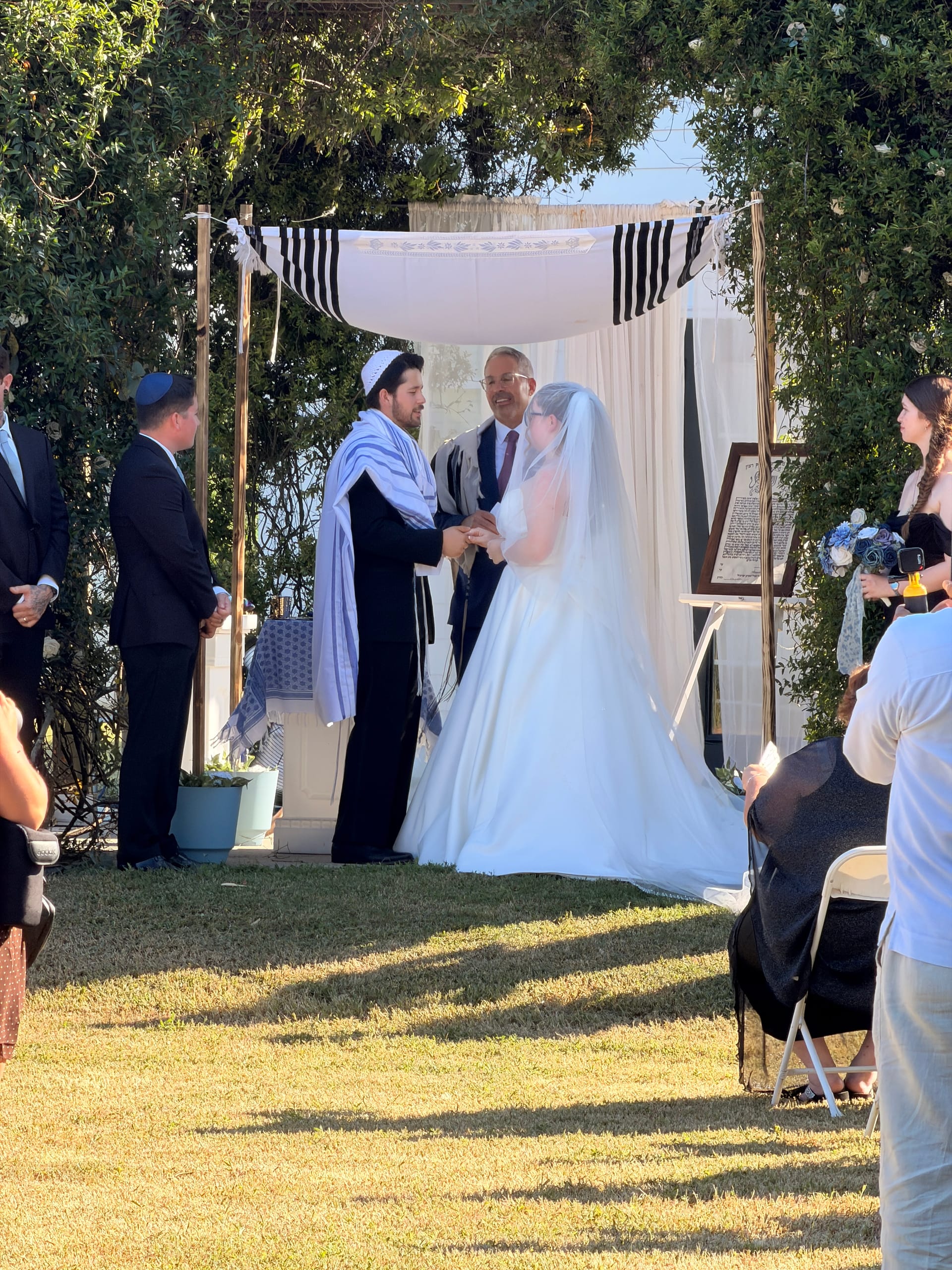 Bride and groom exchange vows beneath a white chuppah adorned with a tallit and draped fabric on the ceremony lawn, surrounded by lush greenery at Scarborough House.