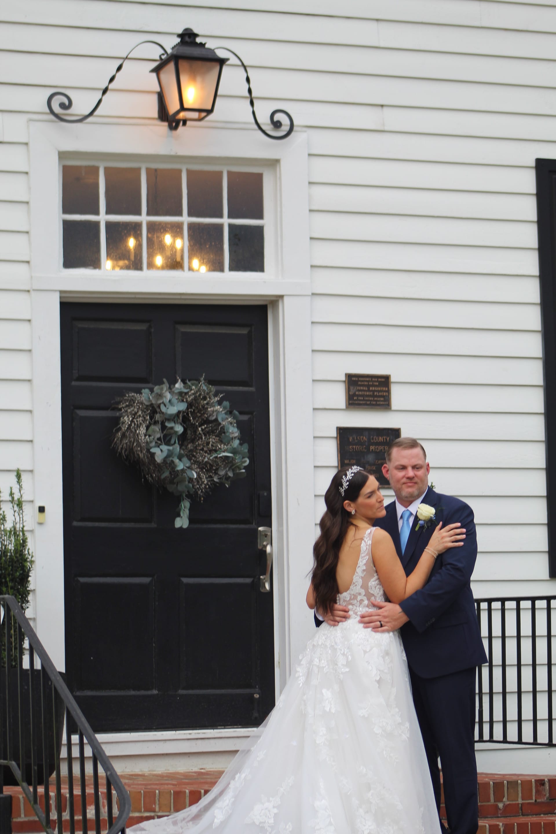 Bride and groom embrace on the front steps of the 1821 Scarborough House manor, framed by a black door with eucalyptus wreath and wrought-iron lantern.