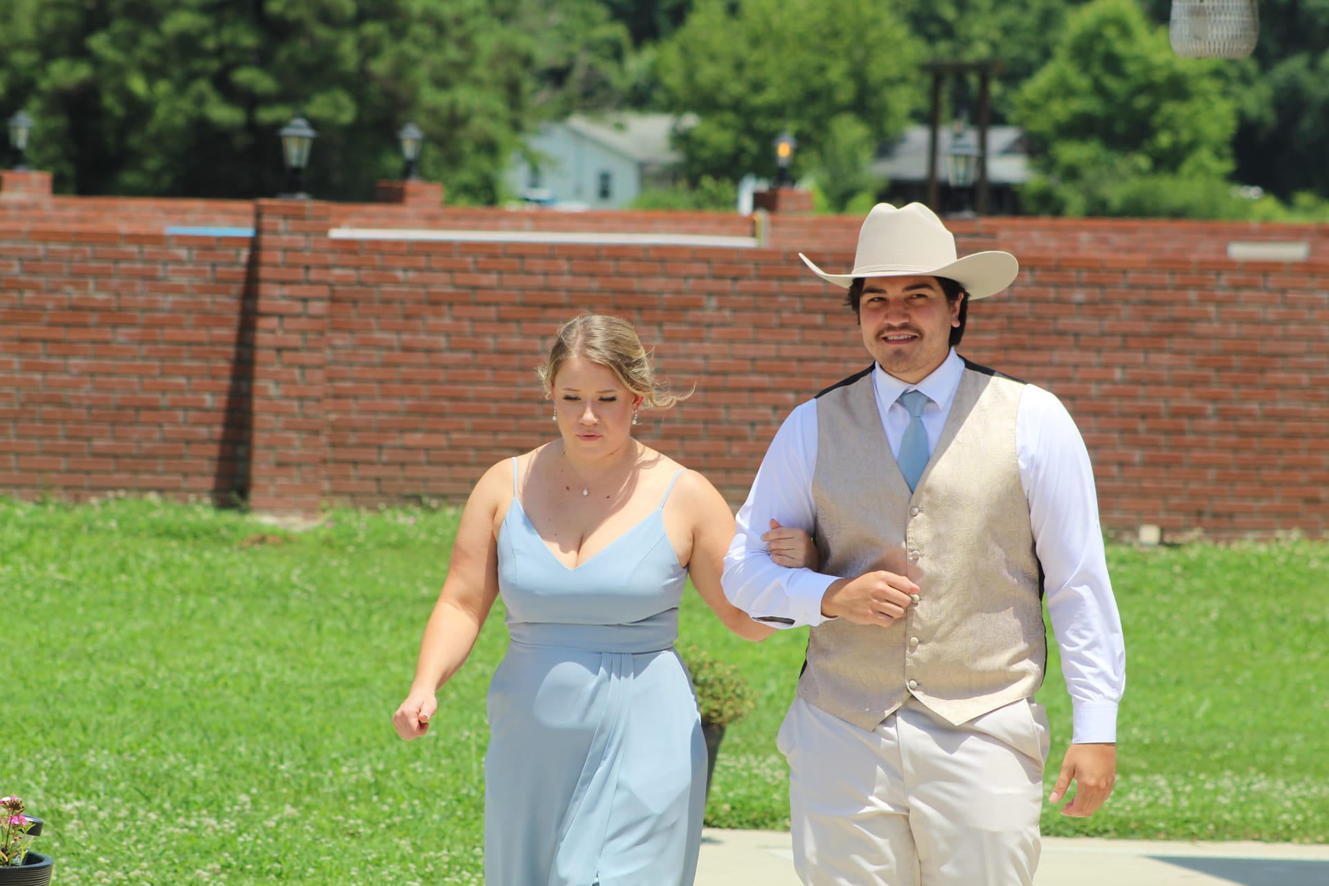 A groomsman in a tan cowboy hat and vest escorts a bridesmaid in a light blue gown across the sunny green lawn at Scarborough House historic estate.