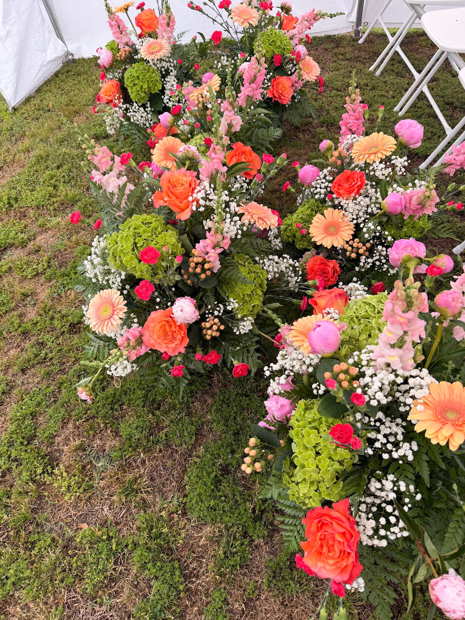 Abundant garden-style floral arrangements featuring orange roses, gerbera daisies, pink snapdragons, green hydrangeas, and baby's breath displayed on the grass at Scarborough House wedding venue.