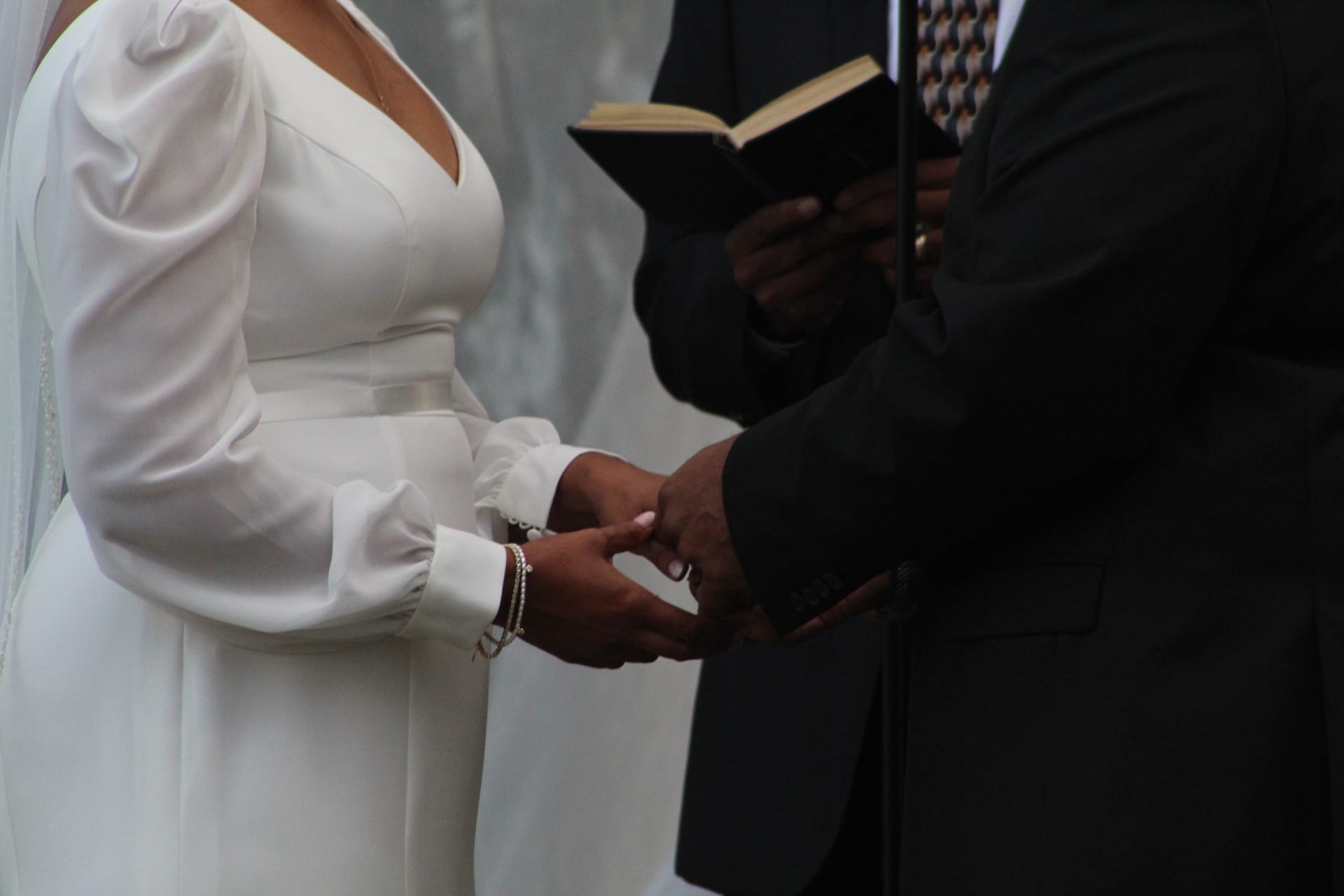 Bride in a long-sleeve white gown holds hands with groom during ring exchange as officiant reads from Bible at Scarborough House outdoor wedding ceremony.