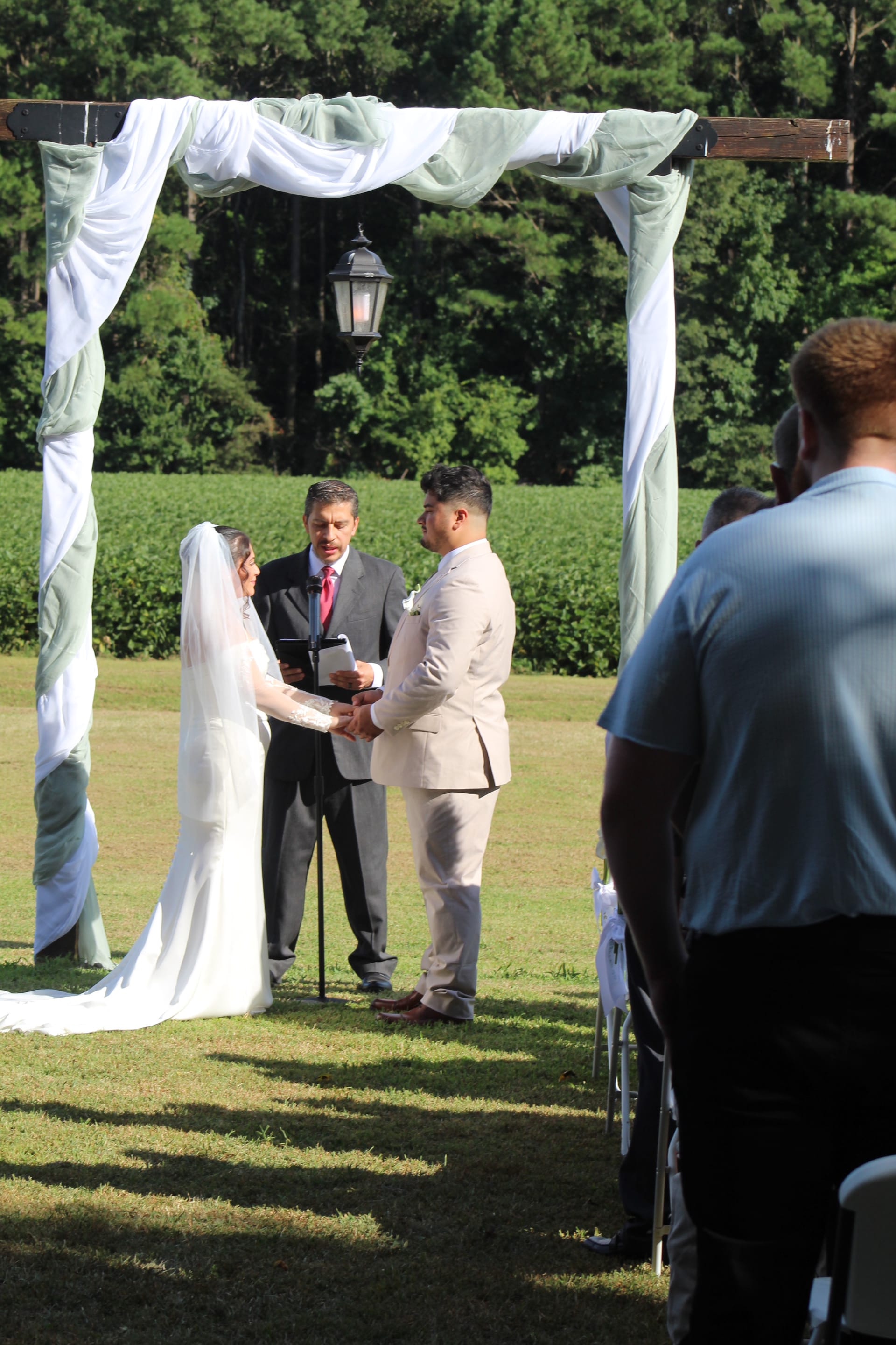 Bride in white gown and veil and groom in tan suit hold hands during outdoor vow exchange beneath a draped wooden arch at Scarborough House estate with green fields in the background.