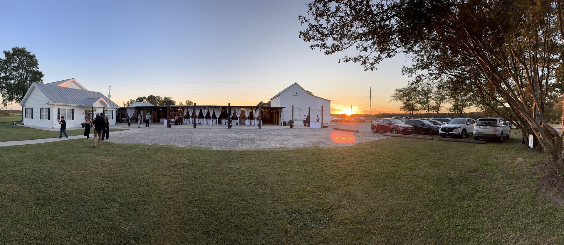 Panoramic golden-hour view of Scarborough House wedding venue at sunset showing the white historic manor, draped magnolia pavilion, and guest-filled courtyard bathed in warm orange light.