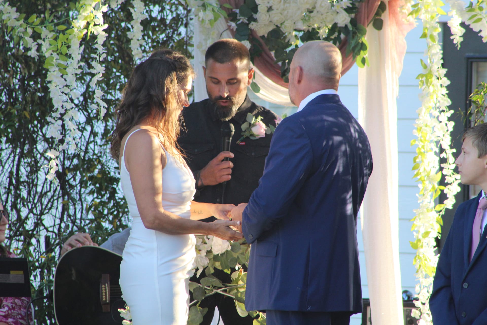 Bride in white gown and groom in navy suit exchange rings during an outdoor wedding ceremony beneath a floral arch adorned with white blooms and greenery at Scarborough House.