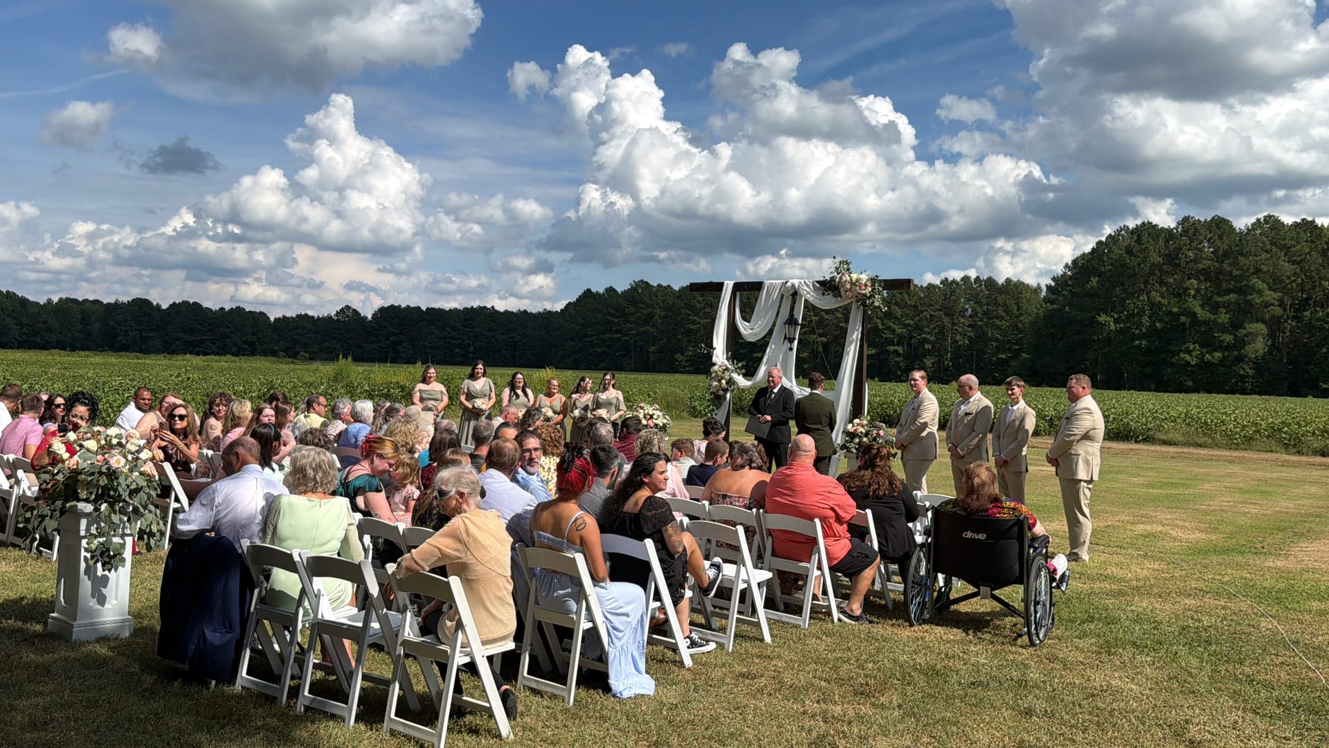 Outdoor wedding ceremony in progress on the Scarborough House ceremony lawn with guests seated in white chairs facing a draped floral arch, bridal party standing alongside, lush farmland fields and dramatic cumulus clouds in the background.
