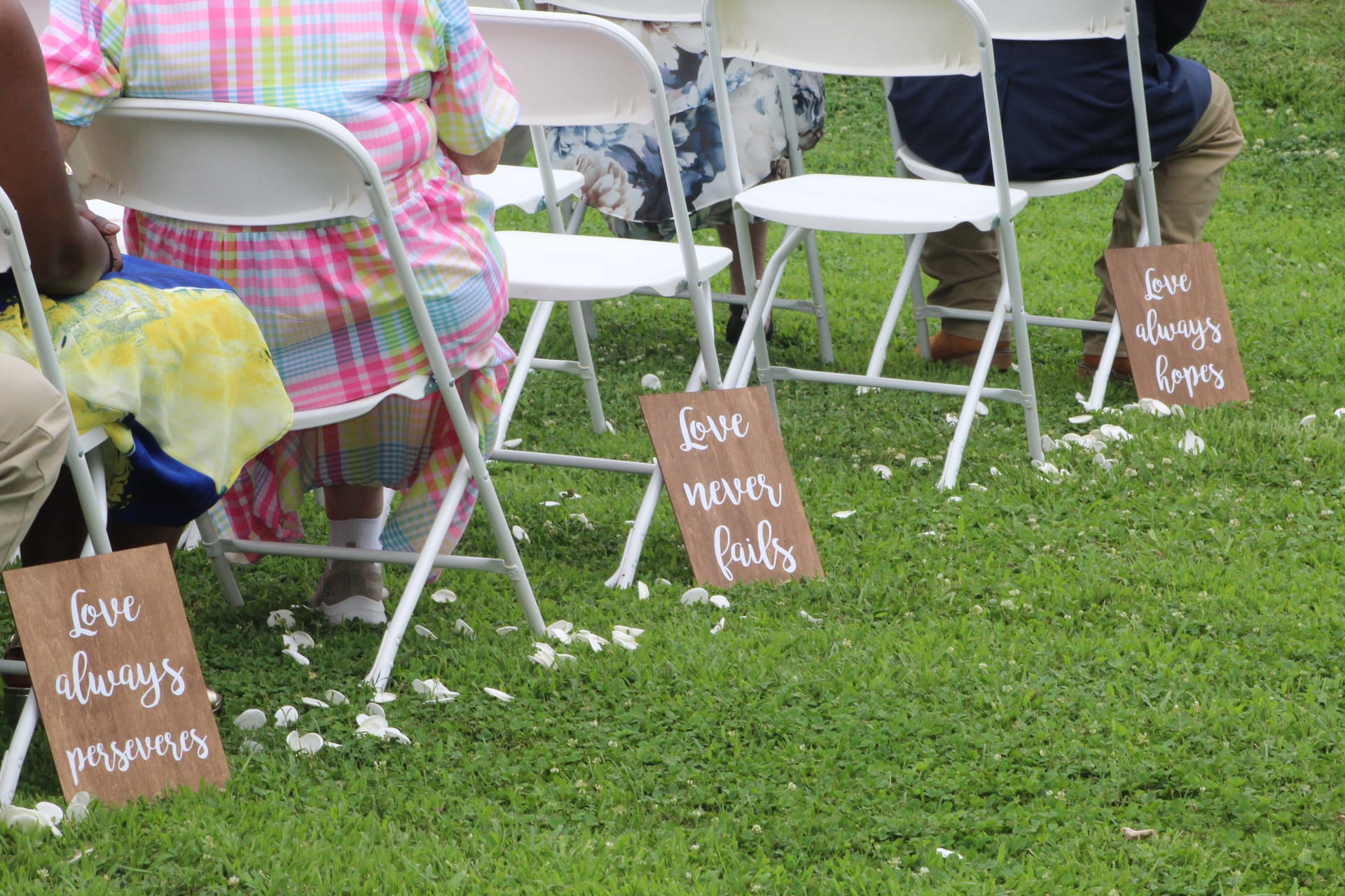 Wooden signs reading Love Always Perseveres, Love Never Fails, and Love Always Hopes line the grass aisle at an outdoor wedding ceremony with white folding chairs and scattered flower petals.