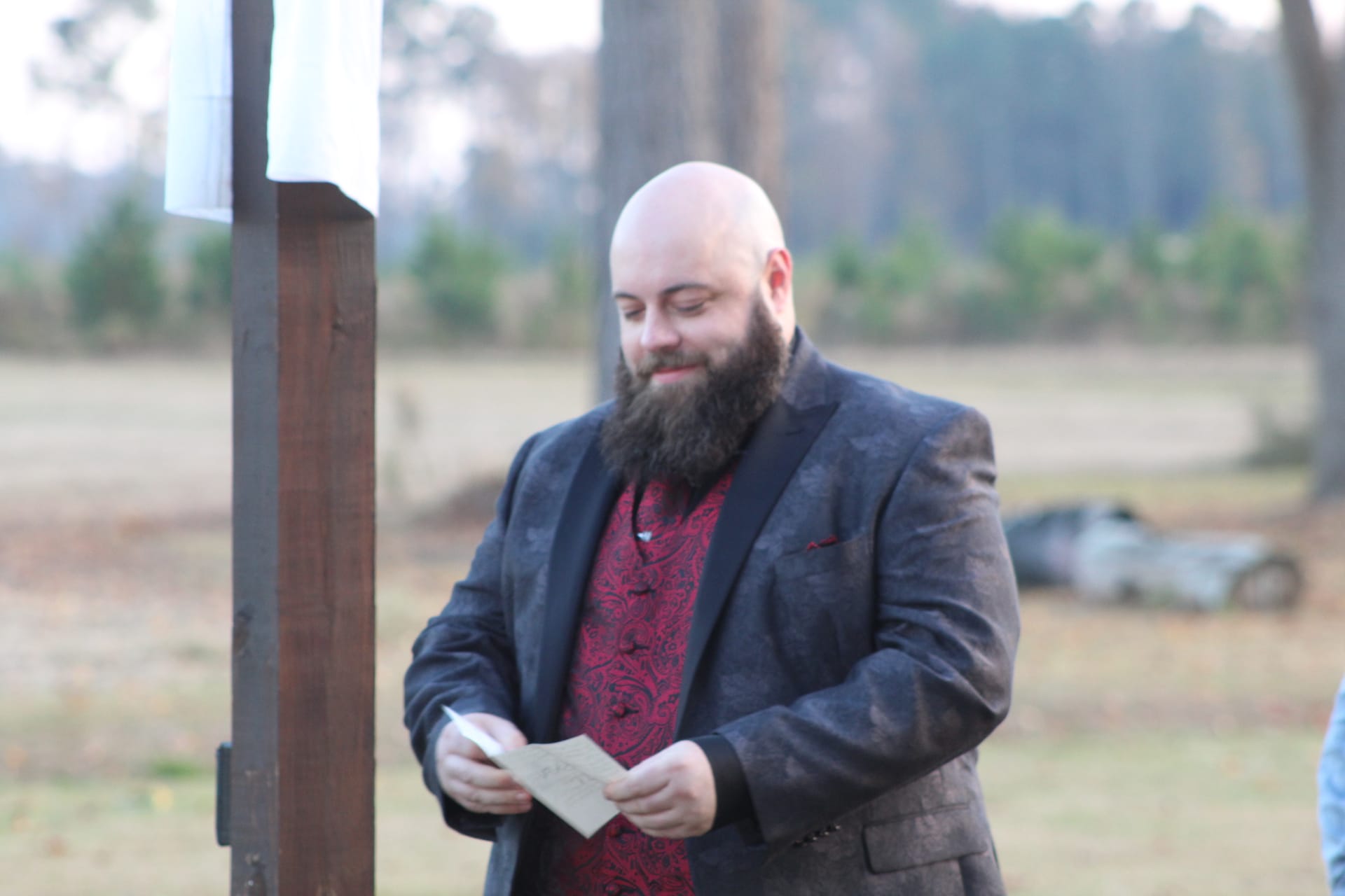 A bald bearded man in a dark blazer and red paisley shirt smiles while reading from a card during an outdoor wedding ceremony at Scarborough House estate.