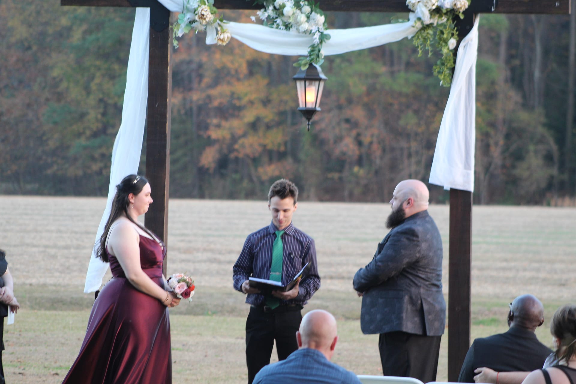 Bride in a burgundy gown holding a bouquet stands before a wood arbor draped in white fabric and white florals as the officiant reads during an outdoor autumn wedding ceremony at Scarborough House.