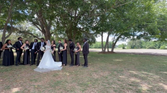Wedding party dressed in black flanking a bride in a white gown and veil gathered beneath large oak trees on the ceremony lawn at Scarborough House.