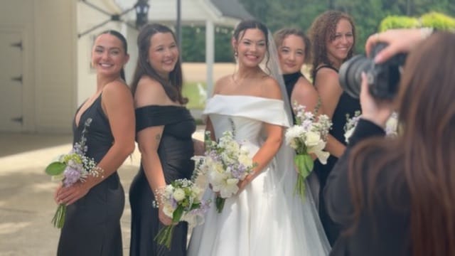 Bride in an off-shoulder white gown poses with four bridesmaids in black dresses holding floral bouquets while a photographer captures the moment outdoors at Scarborough House.