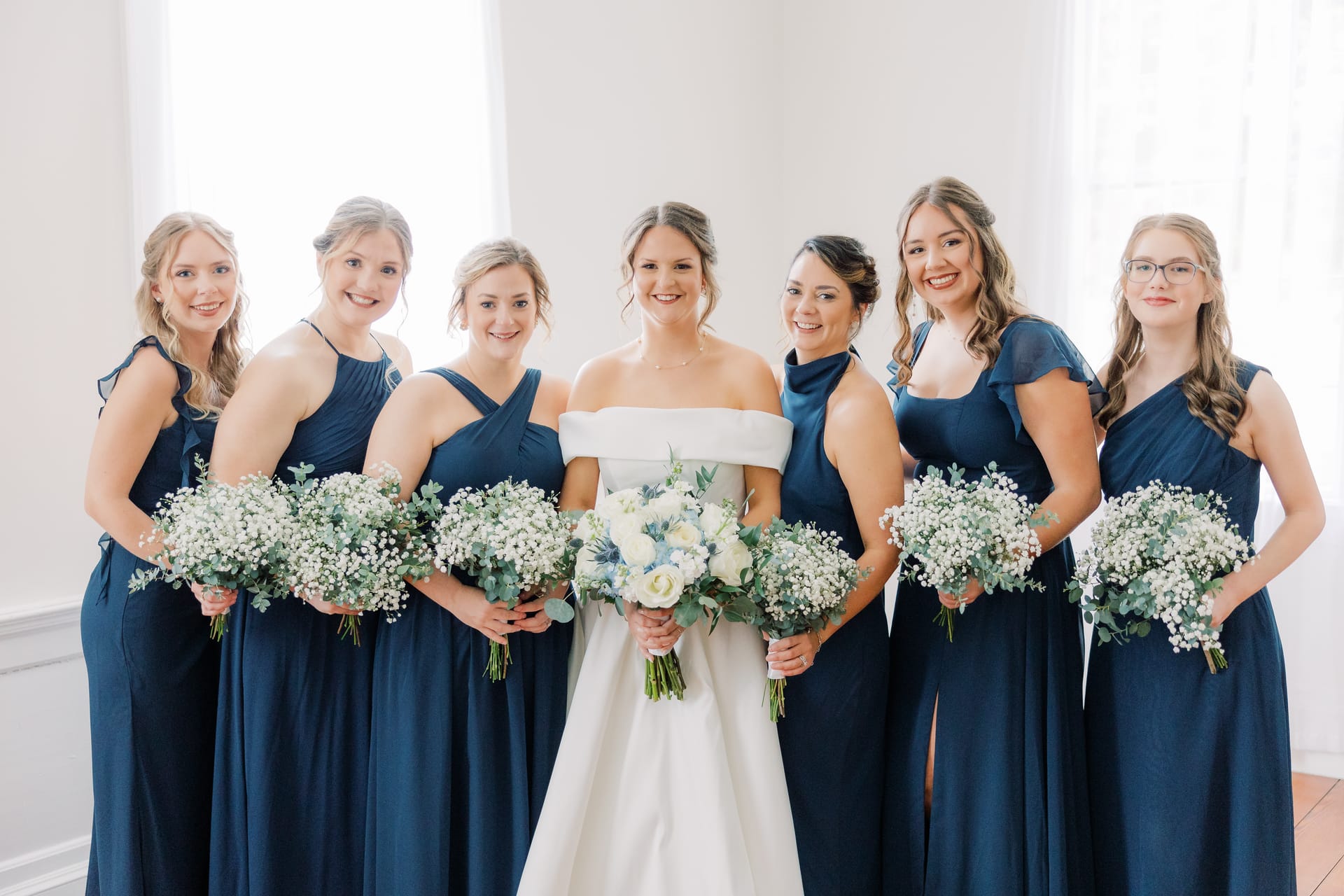 Bride in an off-shoulder white gown stands with six bridesmaids in navy blue dresses holding baby's breath and eucalyptus bouquets inside Scarborough House.