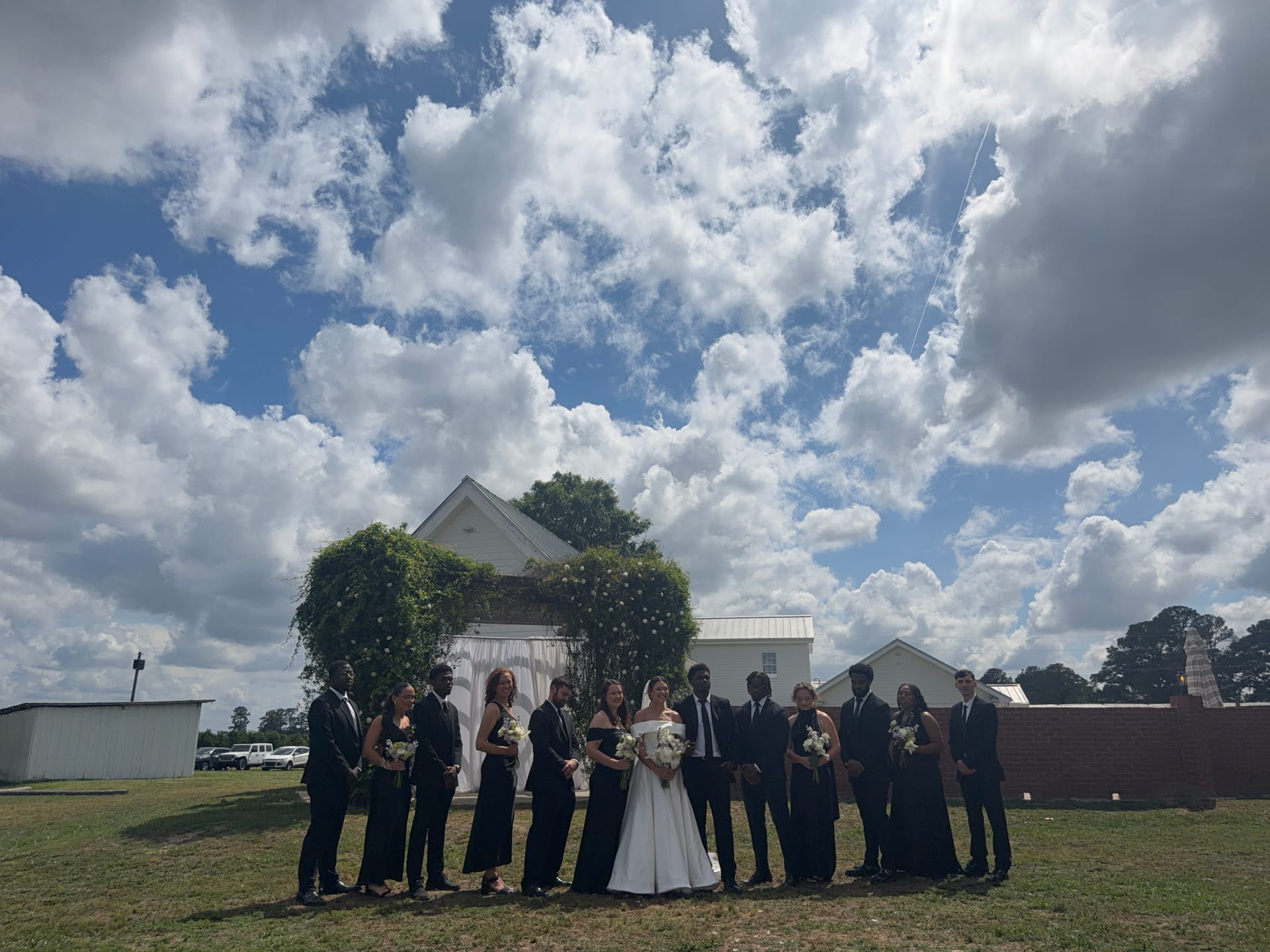 Wedding party dressed in black formal attire poses with the bride in white before a floral archway at Scarborough House under a dramatic blue and cloudy sky.