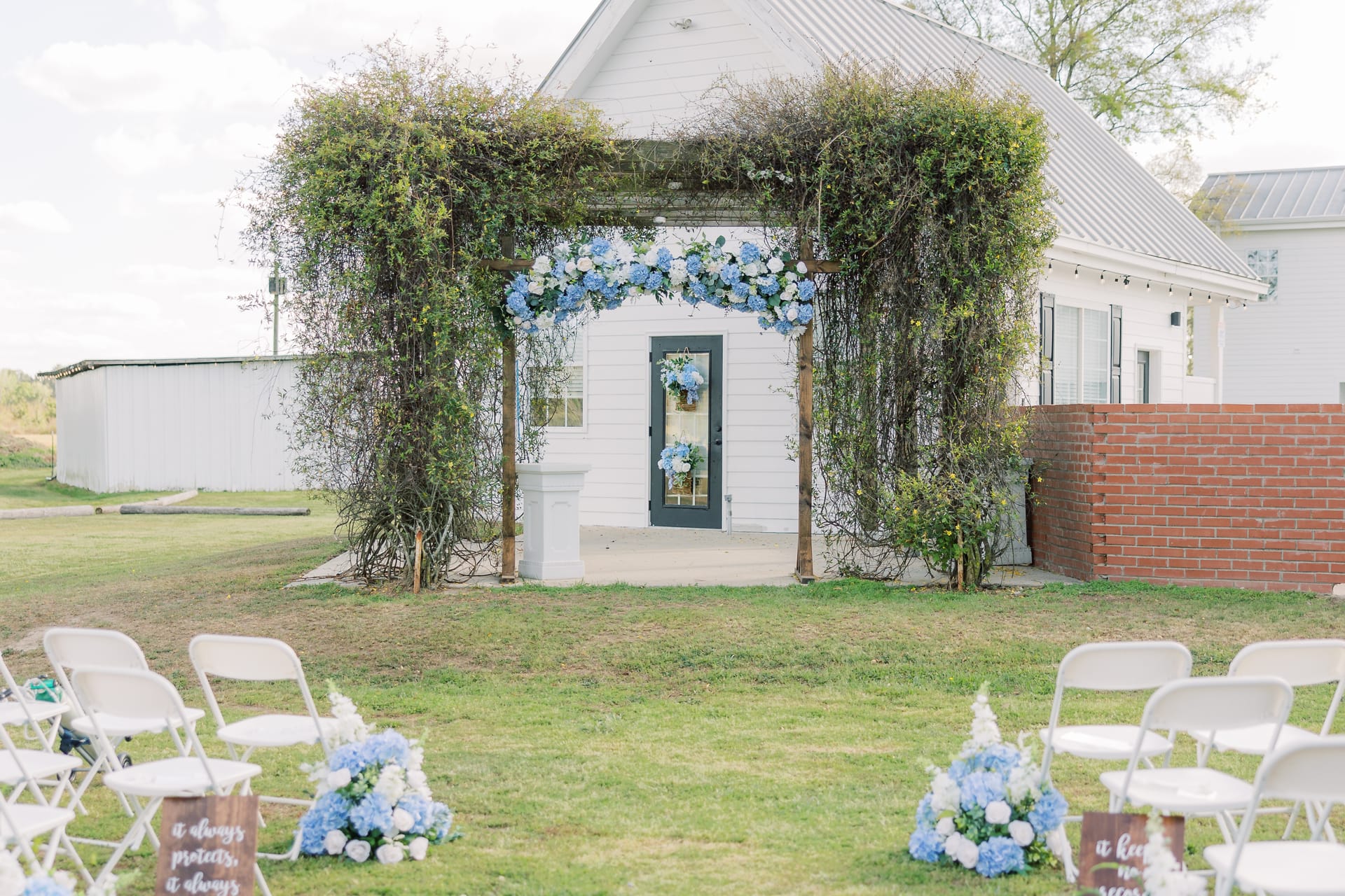Outdoor wedding ceremony lawn at Scarborough House with white folding chairs, blue and white hydrangea arrangements, and a vine-covered archway in front of the chapel door.