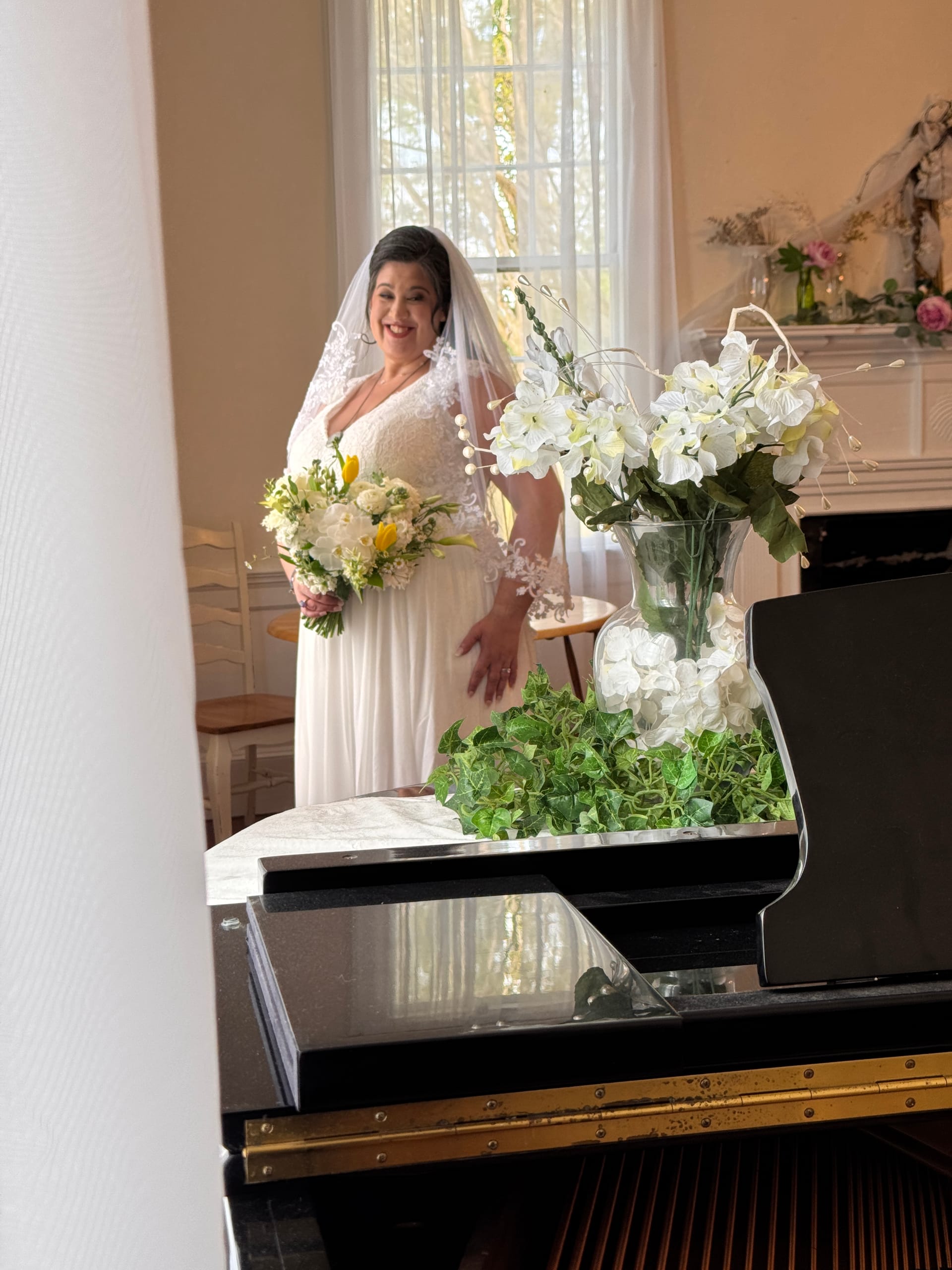 Bride in a white lace gown and veil holding a yellow and white bouquet stands smiling near a window inside the grand hall of Scarborough House historic estate.