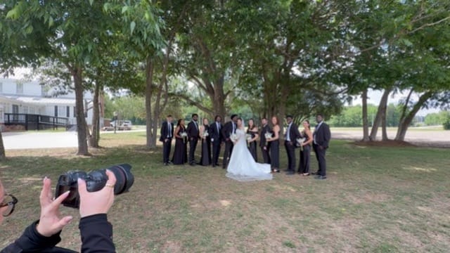 Full wedding party in black attire posing with bride and groom in white under large oak trees at Scarborough House historic estate venue.