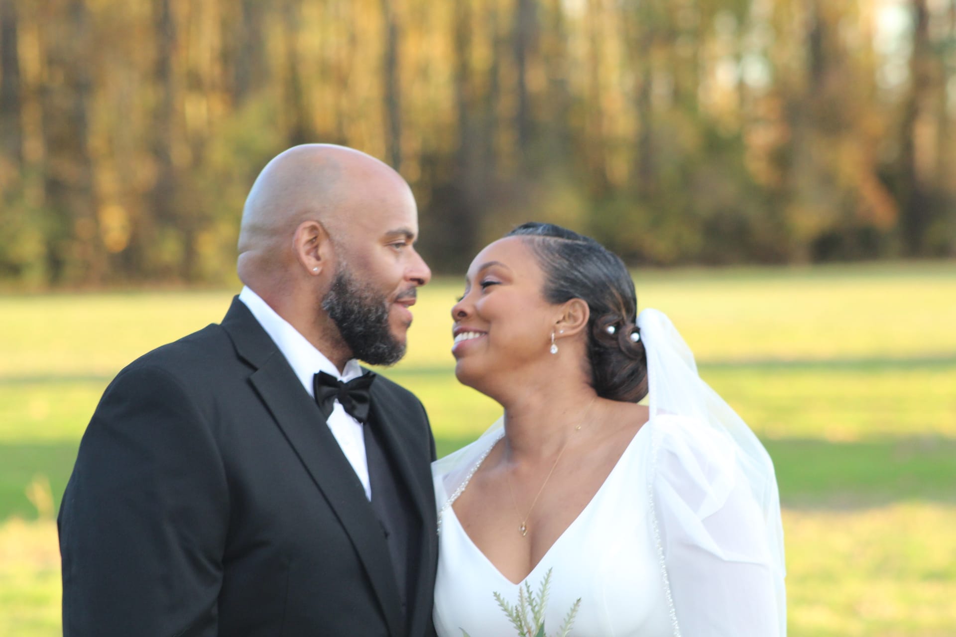 Bride in white gown and groom in black tuxedo gazing lovingly at each other during golden hour with a sunlit field and autumn trees in the background at Scarborough House.