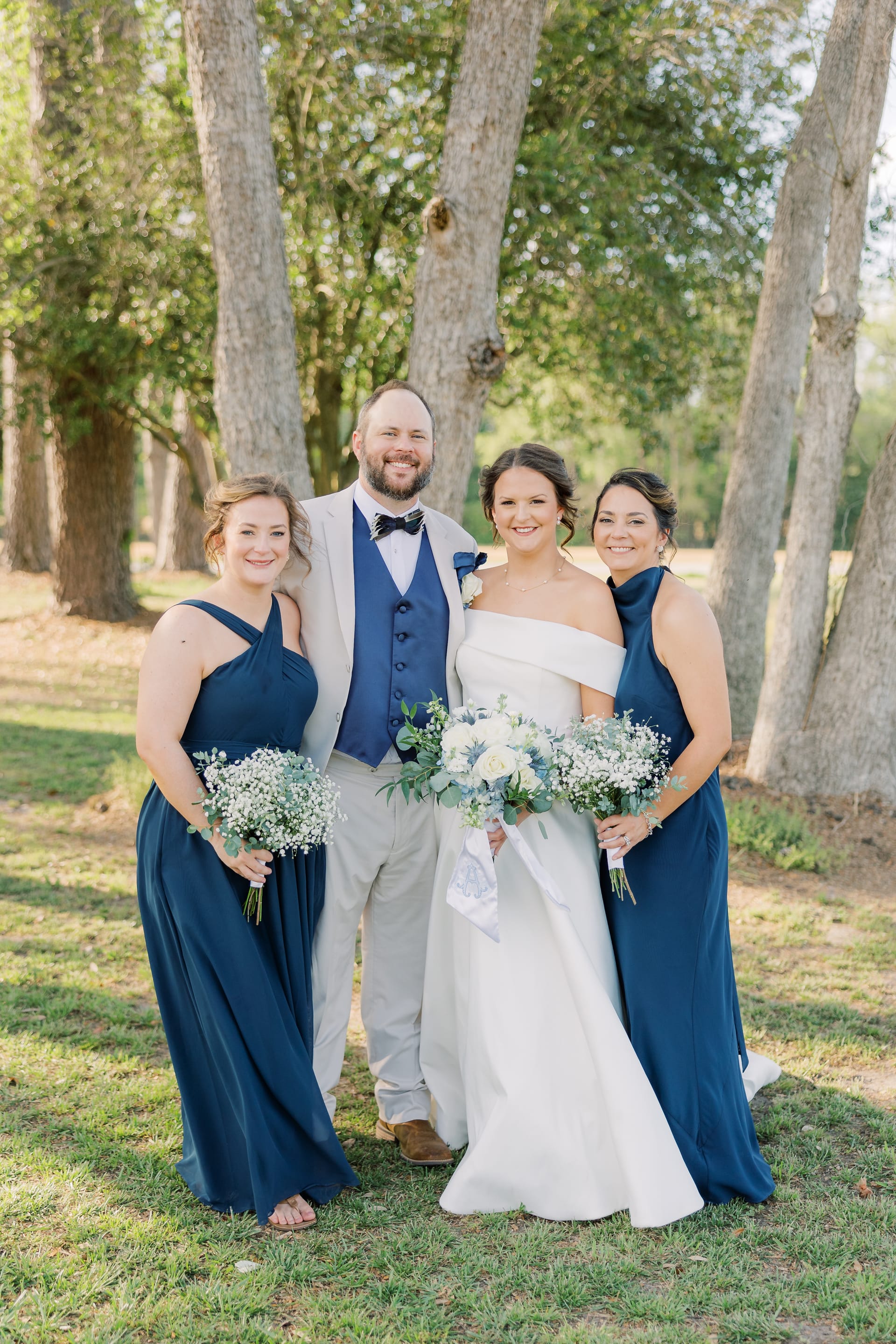 Bride in an off-shoulder white gown and groom in a grey suit with navy vest pose with two bridesmaids in navy dresses holding white bouquets among tall trees at Scarborough House.