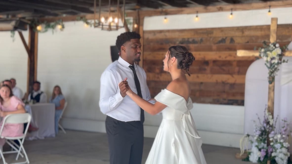 Bride in an off-the-shoulder white gown and groom in a white shirt and dark tie share their first dance in a warmly lit rustic grand hall at Scarborough House as guests look on.