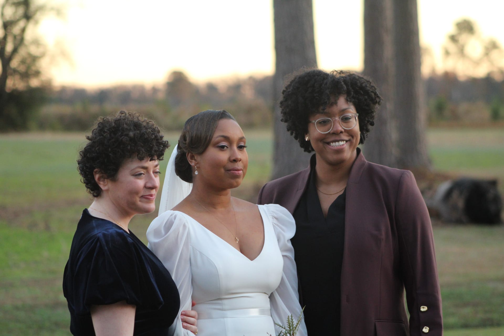 A bride in a white puff-sleeve gown and veil poses with two guests against a golden-hour farmland backdrop at Scarborough House.