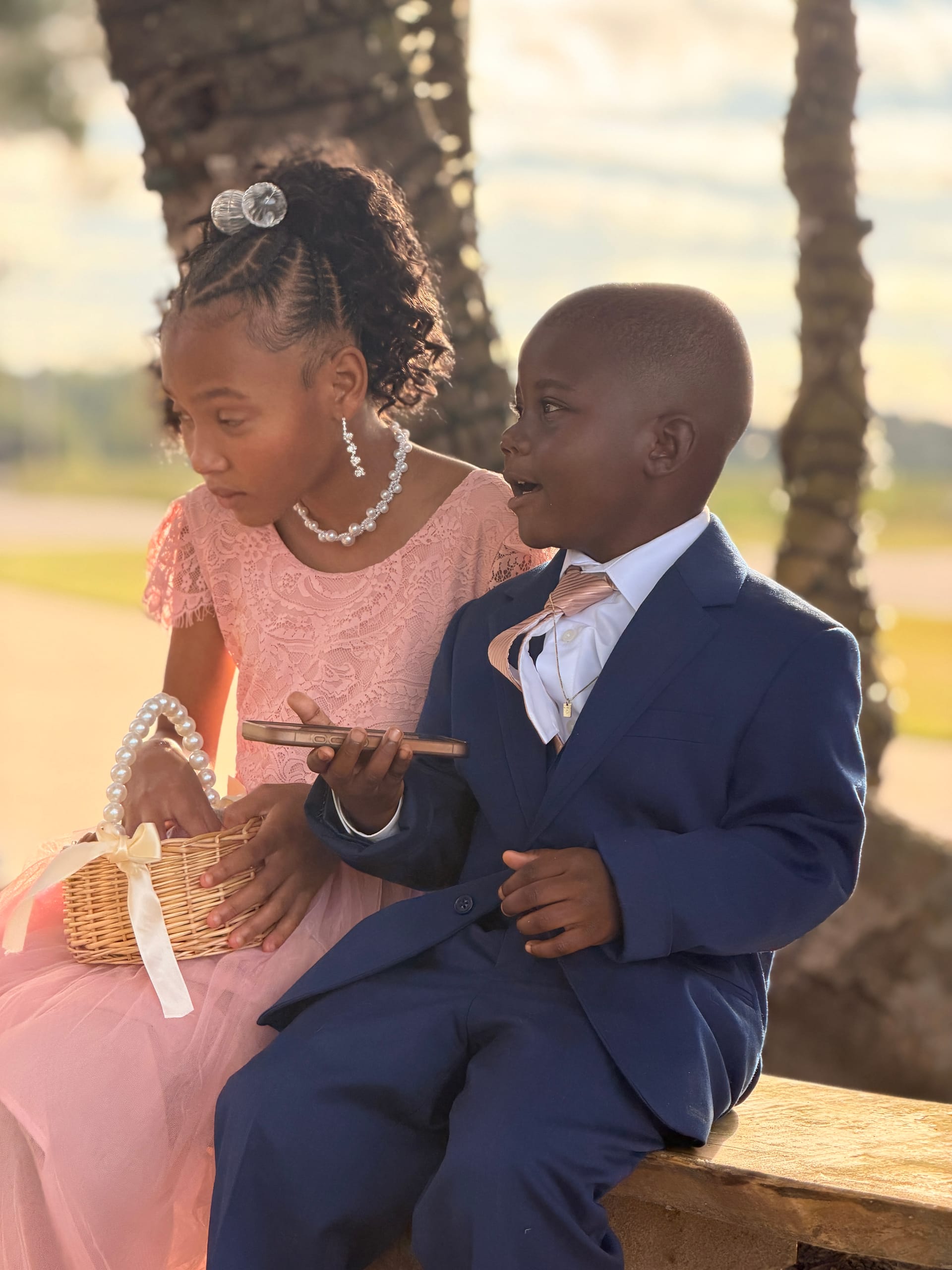 A young flower girl in a blush lace dress and a small ring bearer in a navy suit sit together near palm trees in warm golden-hour light at a wedding celebration.