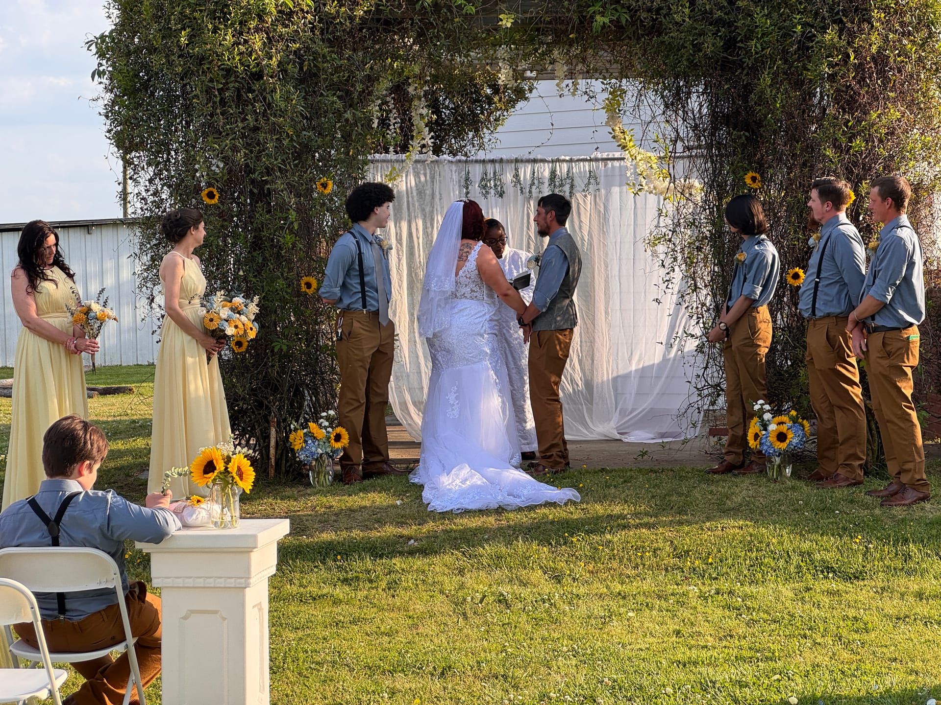 Bride and groom exchange vows beneath a lush greenery arch adorned with sunflowers while bridesmaids in yellow dresses and groomsmen in blue shirts with suspenders look on during an outdoor wedding ceremony.