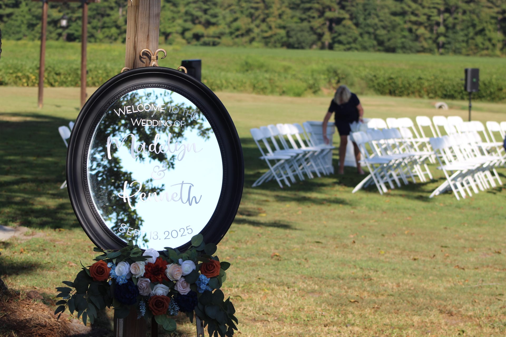 Round black-framed mirror welcome sign reading Welcome to the Wedding of Madelyn and Kenneth September 13 2025 adorned with red white and blue florals at an outdoor ceremony lawn with white folding chairs.