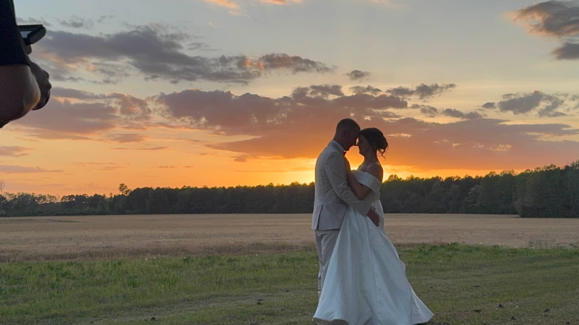 Bride and groom share an intimate forehead-to-forehead moment silhouetted against a dramatic orange and gold sunset over the open farmland fields at Scarborough House.