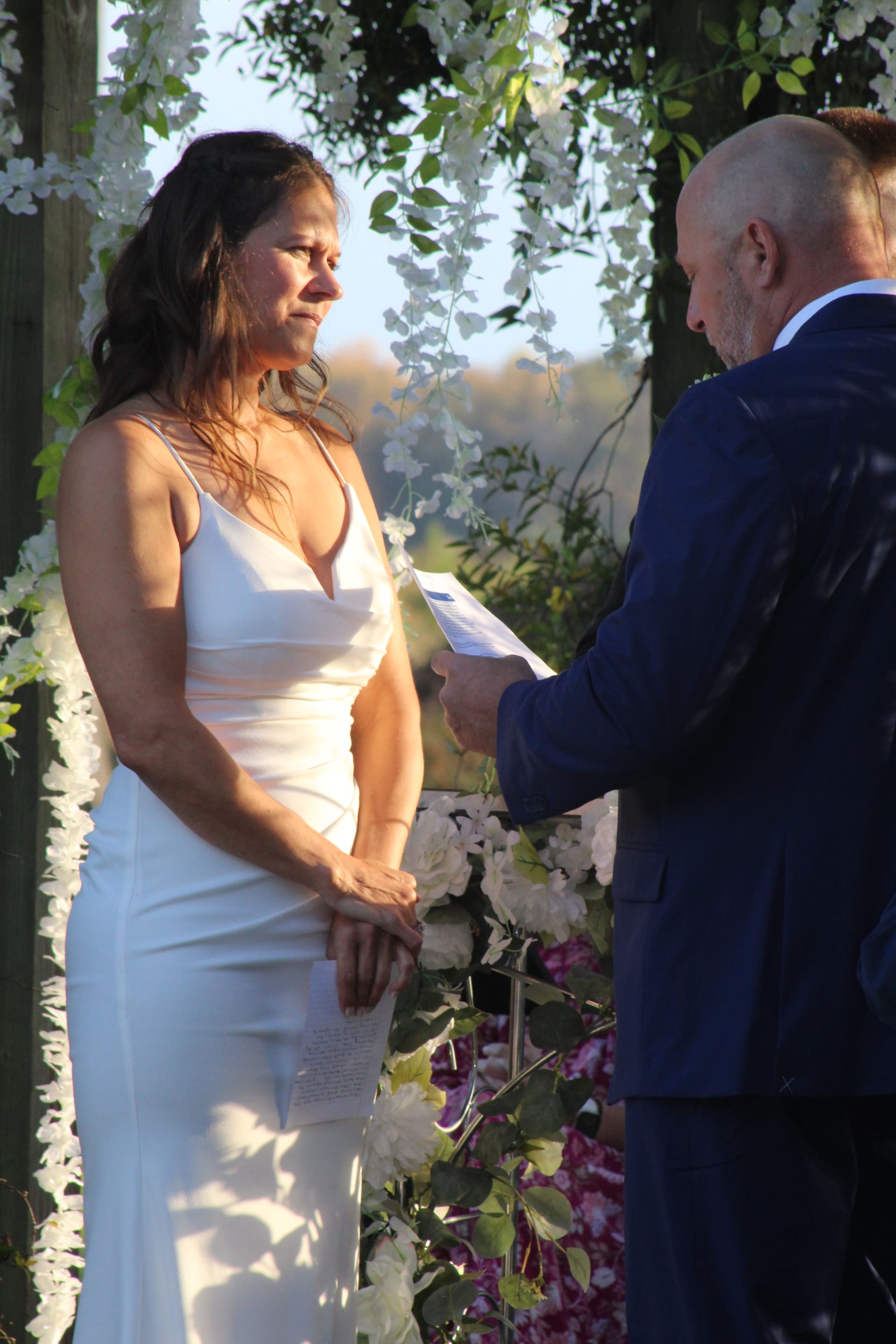 Bride in a sleek white gown listens emotionally as the groom reads his vows beneath a floral arch at golden hour at Scarborough House.
