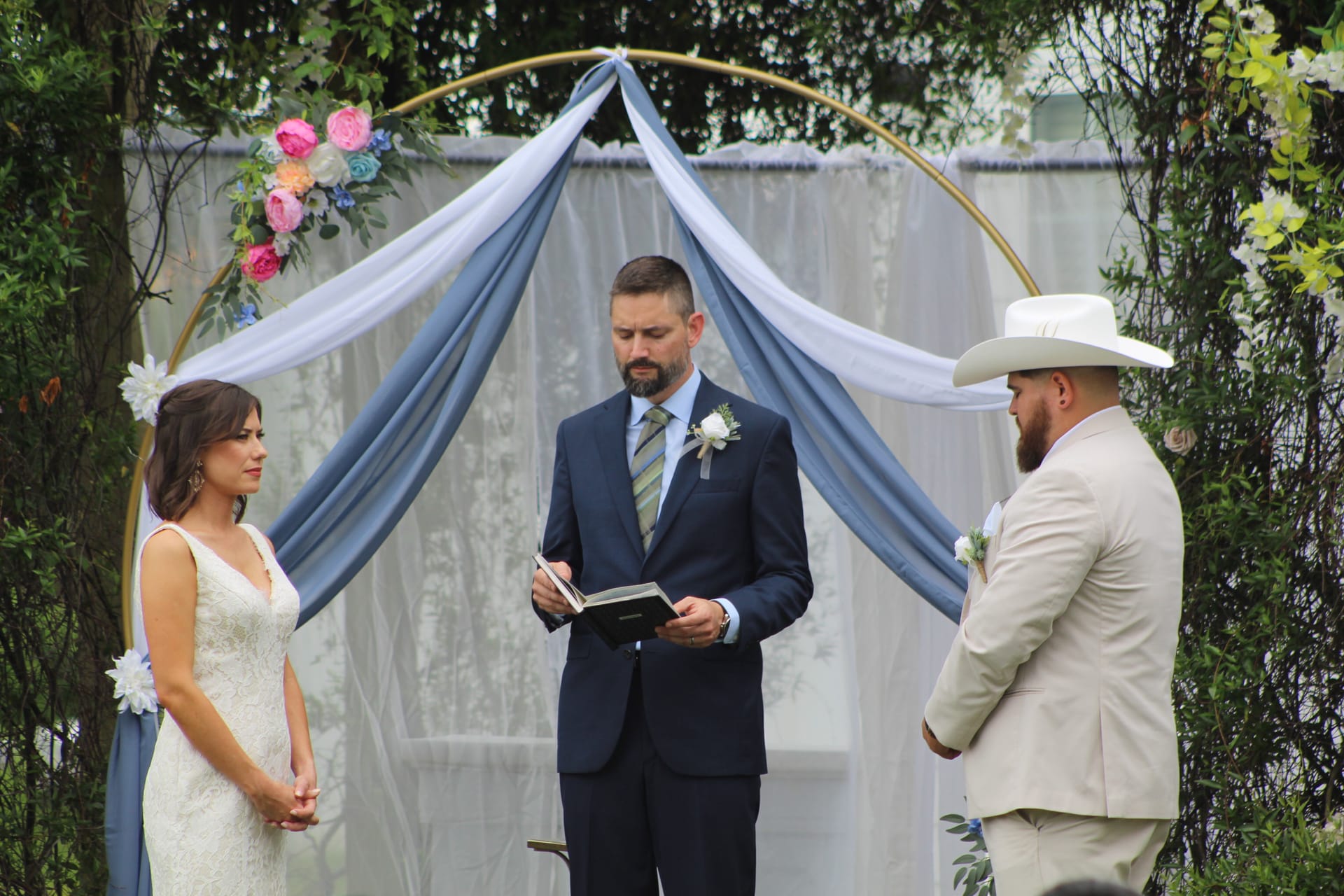Bride in white lace gown and groom in tan suit with white cowboy hat exchange vows beneath a gold circular arch adorned with blue draping and colorful flowers at Scarborough House outdoor ceremony.
