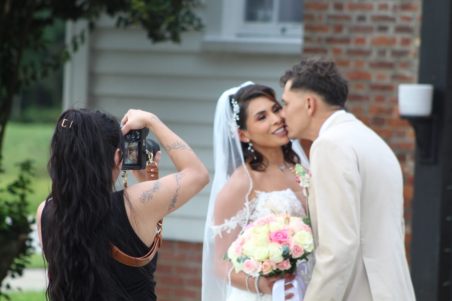 A wedding photographer with floral arm tattoos captures a groom kissing his brides cheek as she smiles and holds a bouquet of pink and white roses in front of a historic brick and wood-sided manor house.