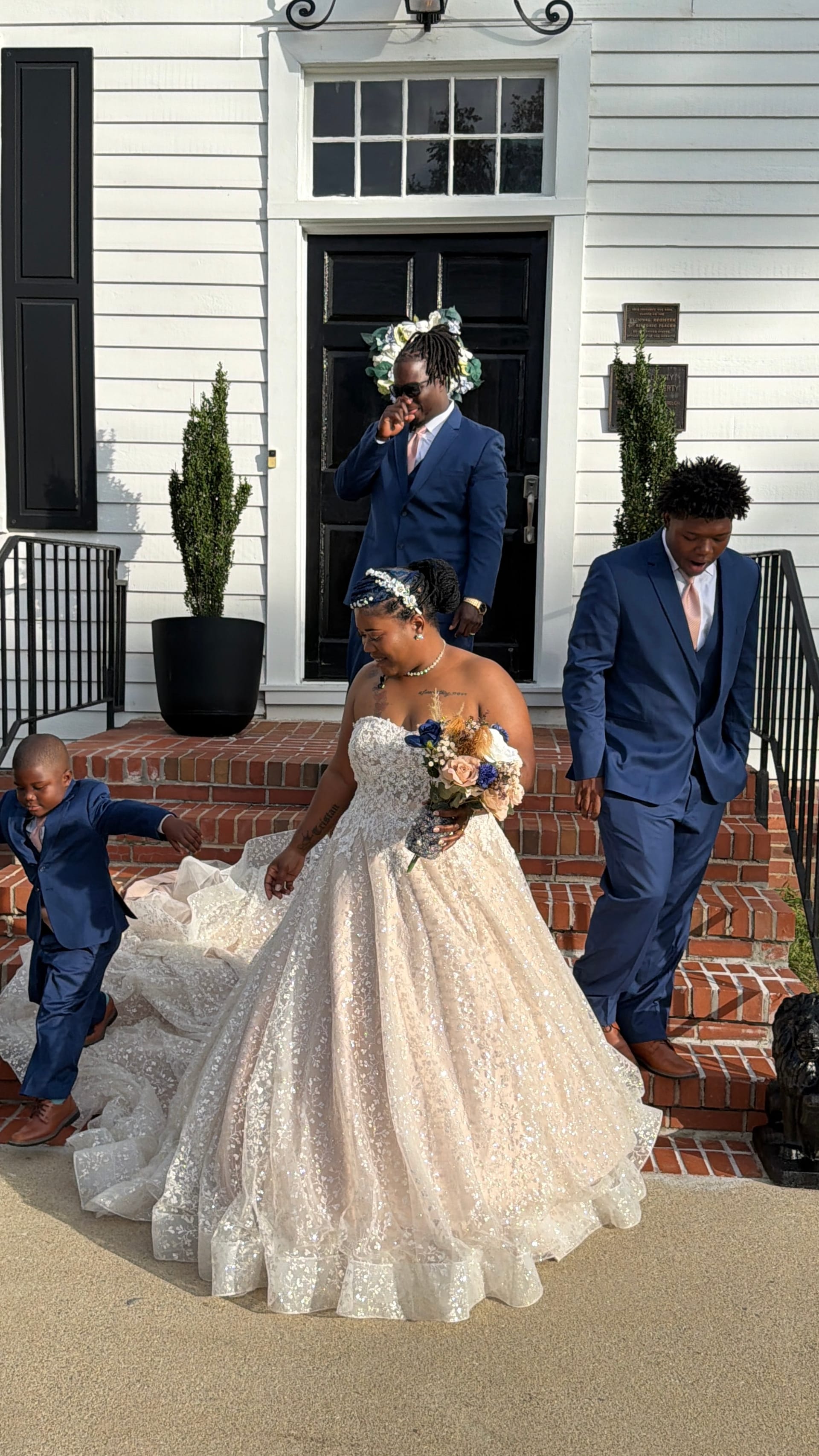 A bride in a voluminous lace ball gown holds her bouquet while descending the brick front steps of the historic 1821 Scarborough House manor, assisted by two young men in navy suits and a small boy in matching attire.