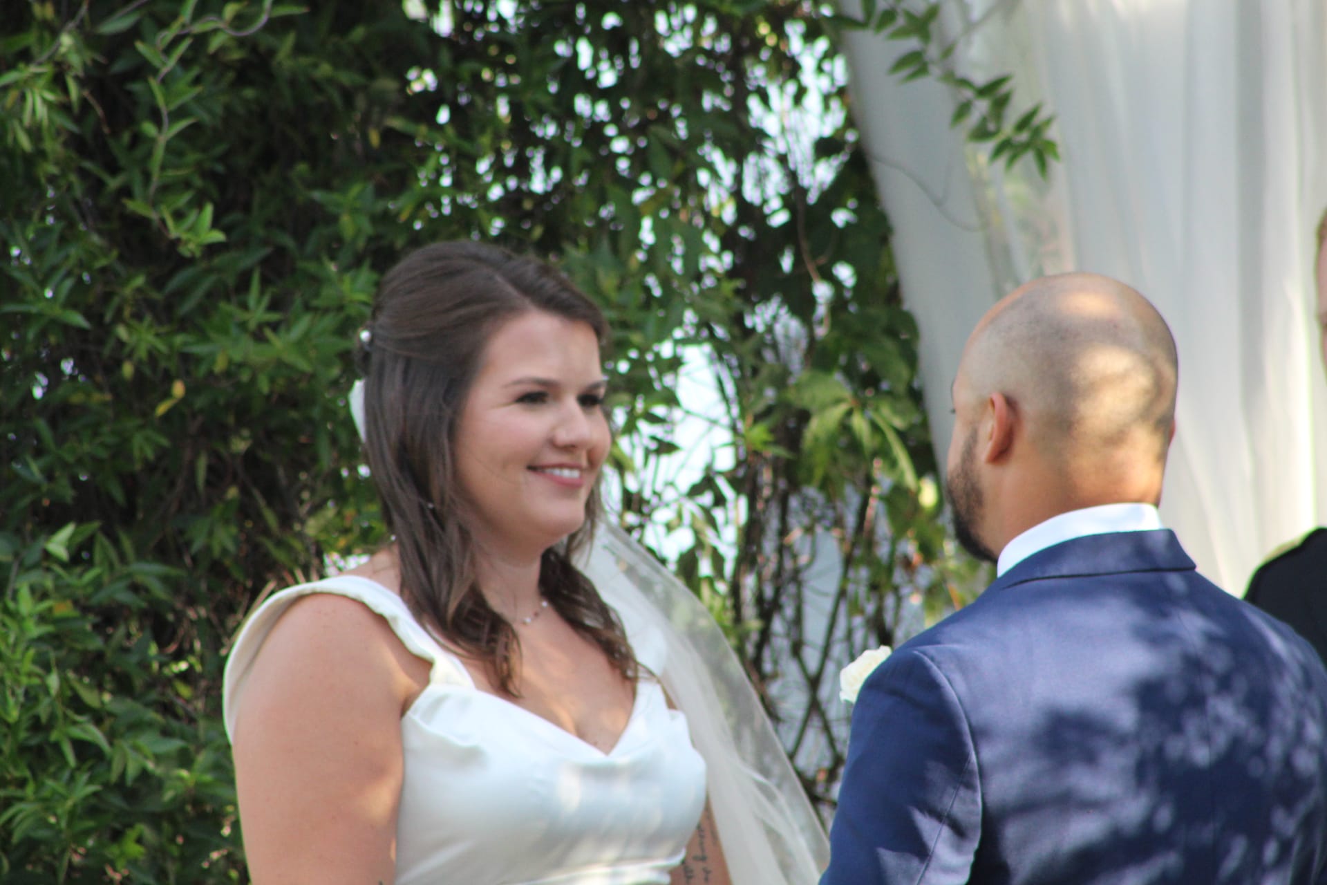 Smiling bride in a white gown faces her groom during an outdoor wedding ceremony surrounded by lush greenery at a historic estate venue.