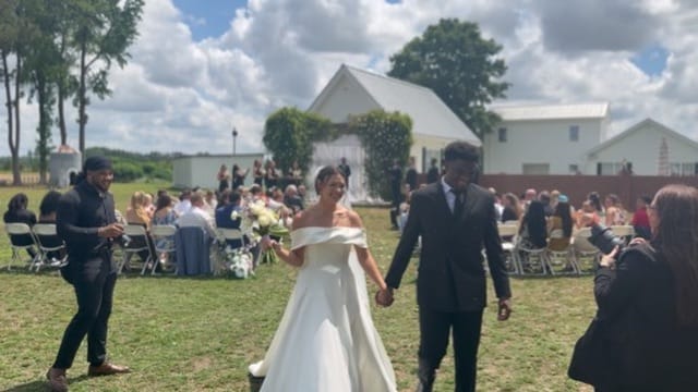 Bride in an off-shoulder white gown and groom in a dark suit walk hand-in-hand down the ceremony lawn aisle at Scarborough House, with seated guests and a white barn backdrop under a partly cloudy summer sky.