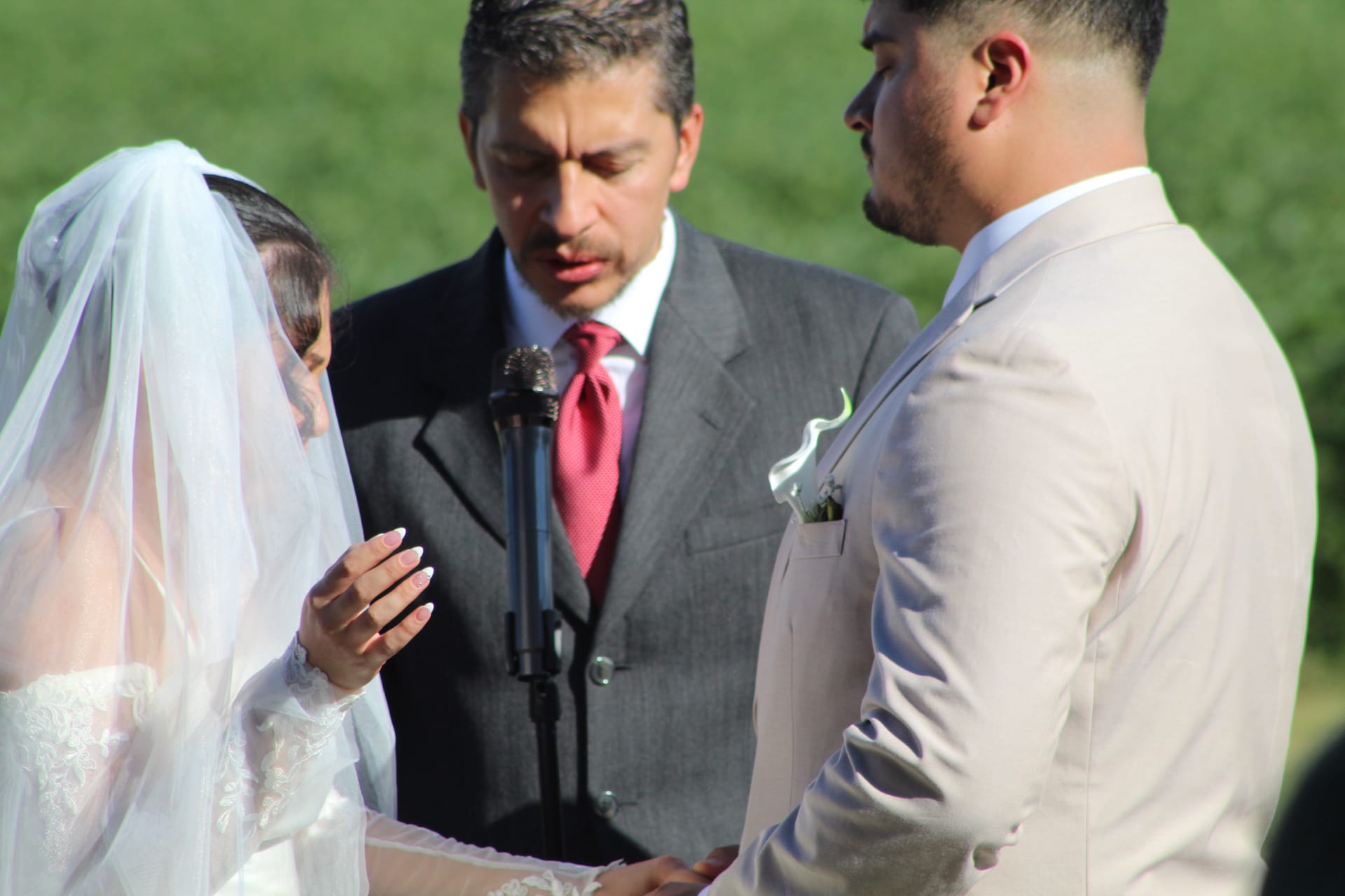 Bride in lace gown and veil exchanges vows with groom in a cream suit while an officiant in a gray suit reads from a book during an outdoor ceremony at Scarborough House.