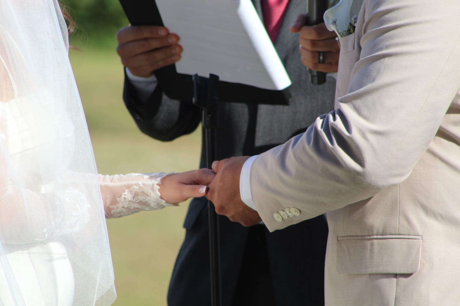 Groom placing a wedding ring on the bride's lace-sleeved hand during an outdoor ceremony at Scarborough House while the officiant reads from a binder in the background.
