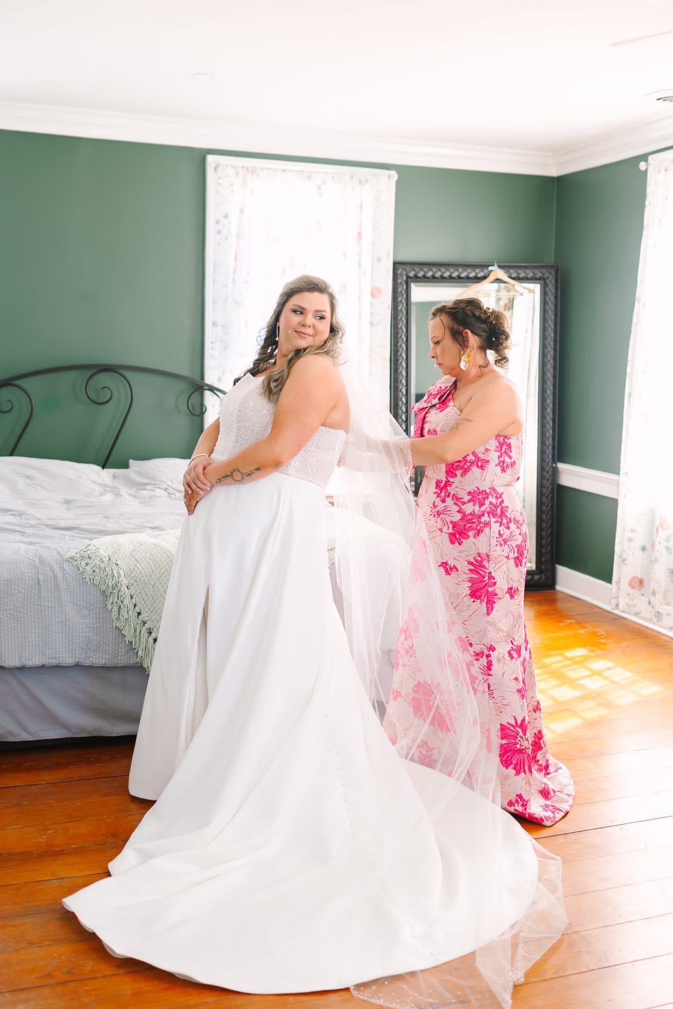 Bride in a flowing white gown looks back with a smile as her attendant in a pink floral dress adjusts her veil in an elegantly appointed green-walled bridal suite at Scarborough House.