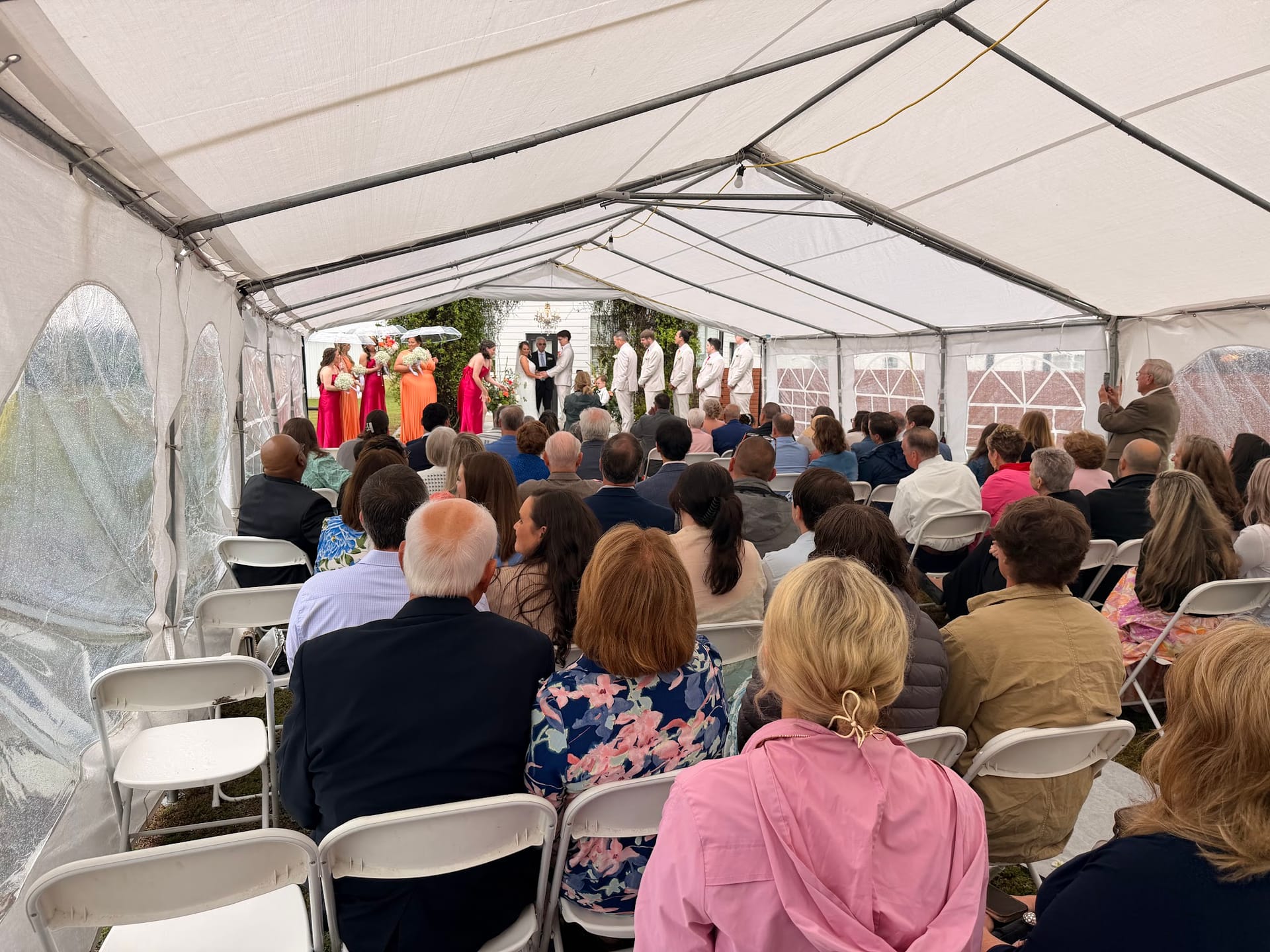 Guests seated in white folding chairs under a white marquee tent watch a wedding ceremony with a colorful bridal party gathered at the altar.
