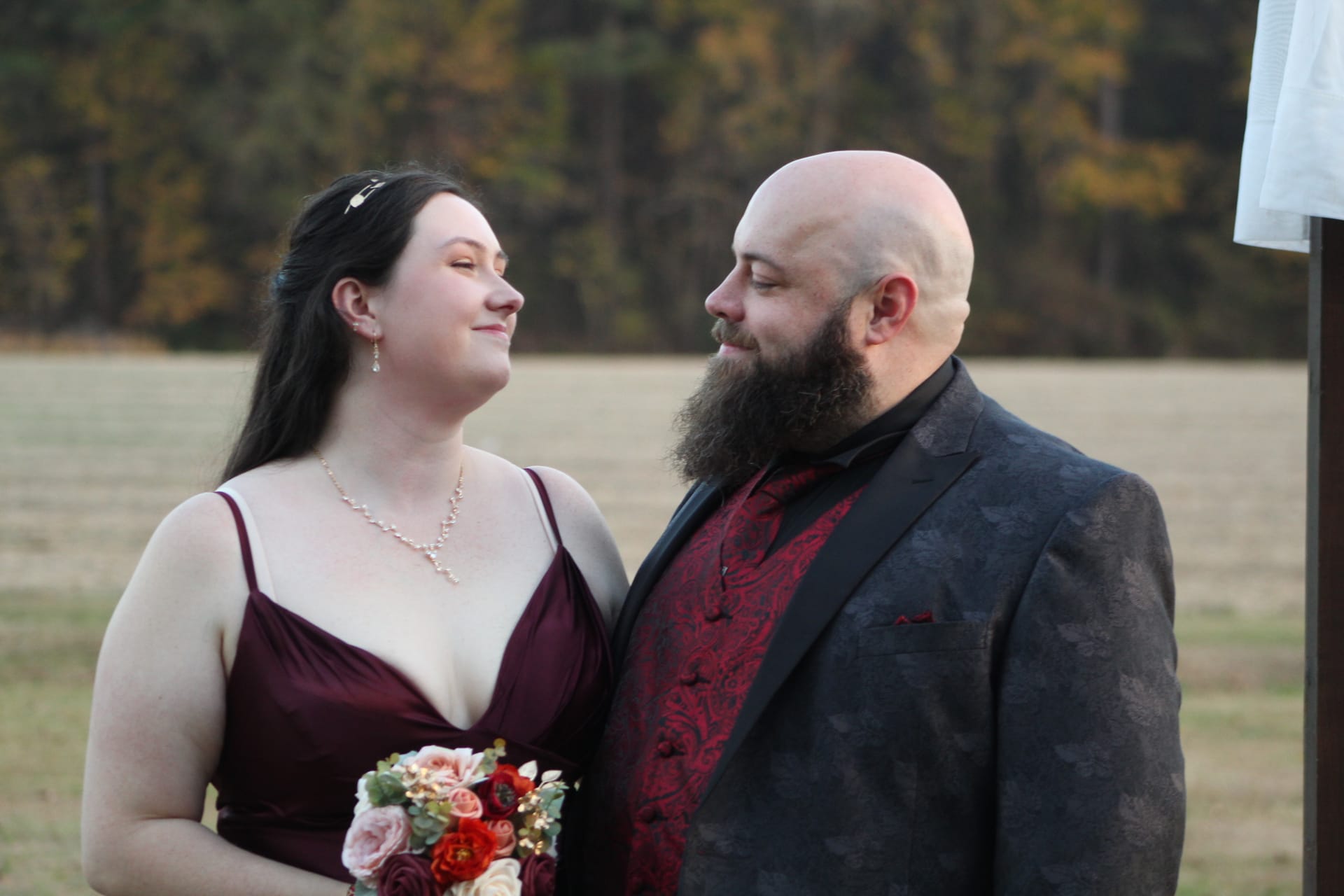 Bride in a burgundy gown holding a floral bouquet gazes lovingly at her bearded groom in a dark patterned suit with a red paisley vest during an outdoor ceremony at Scarborough House estate.
