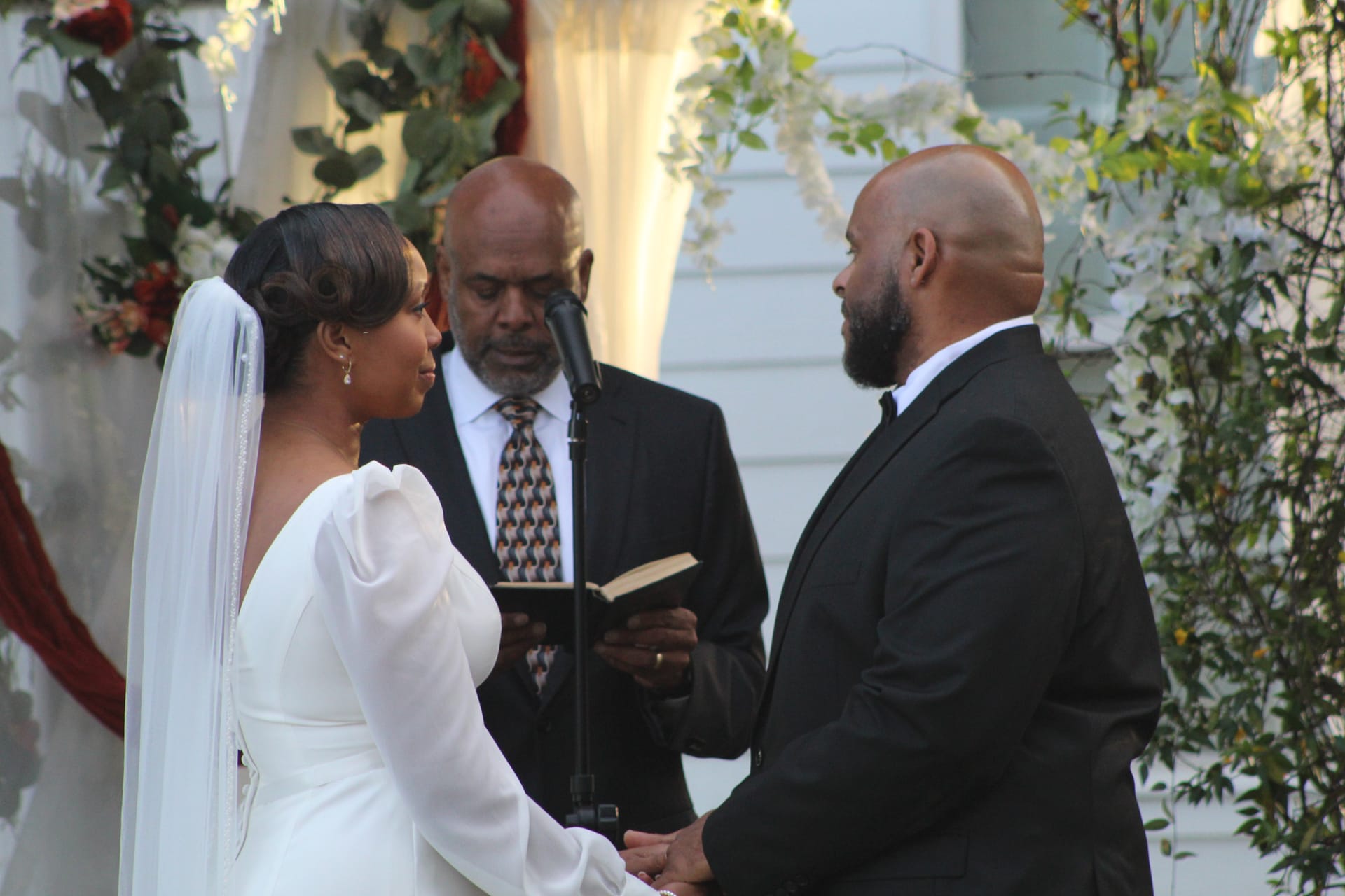 Bride in a long white gown and veil and groom in a black suit hold hands and face each other during their outdoor wedding vow exchange at Scarborough House while an officiant reads from a book beneath a floral arch.