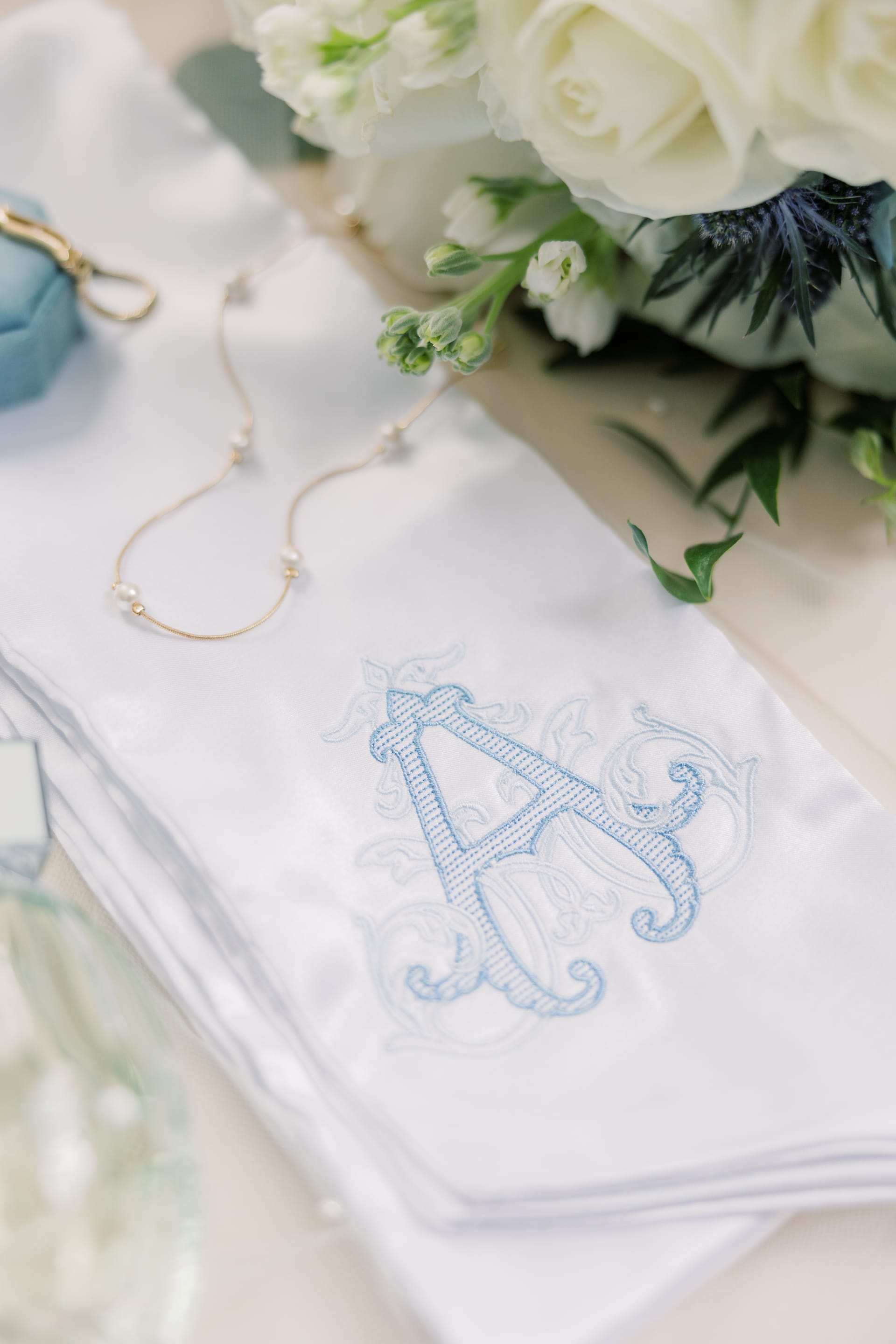 A monogrammed white handkerchief embroidered with a blue decorative letter A rests beside a delicate pearl necklace and white floral bouquet with thistle accents at Scarborough House.