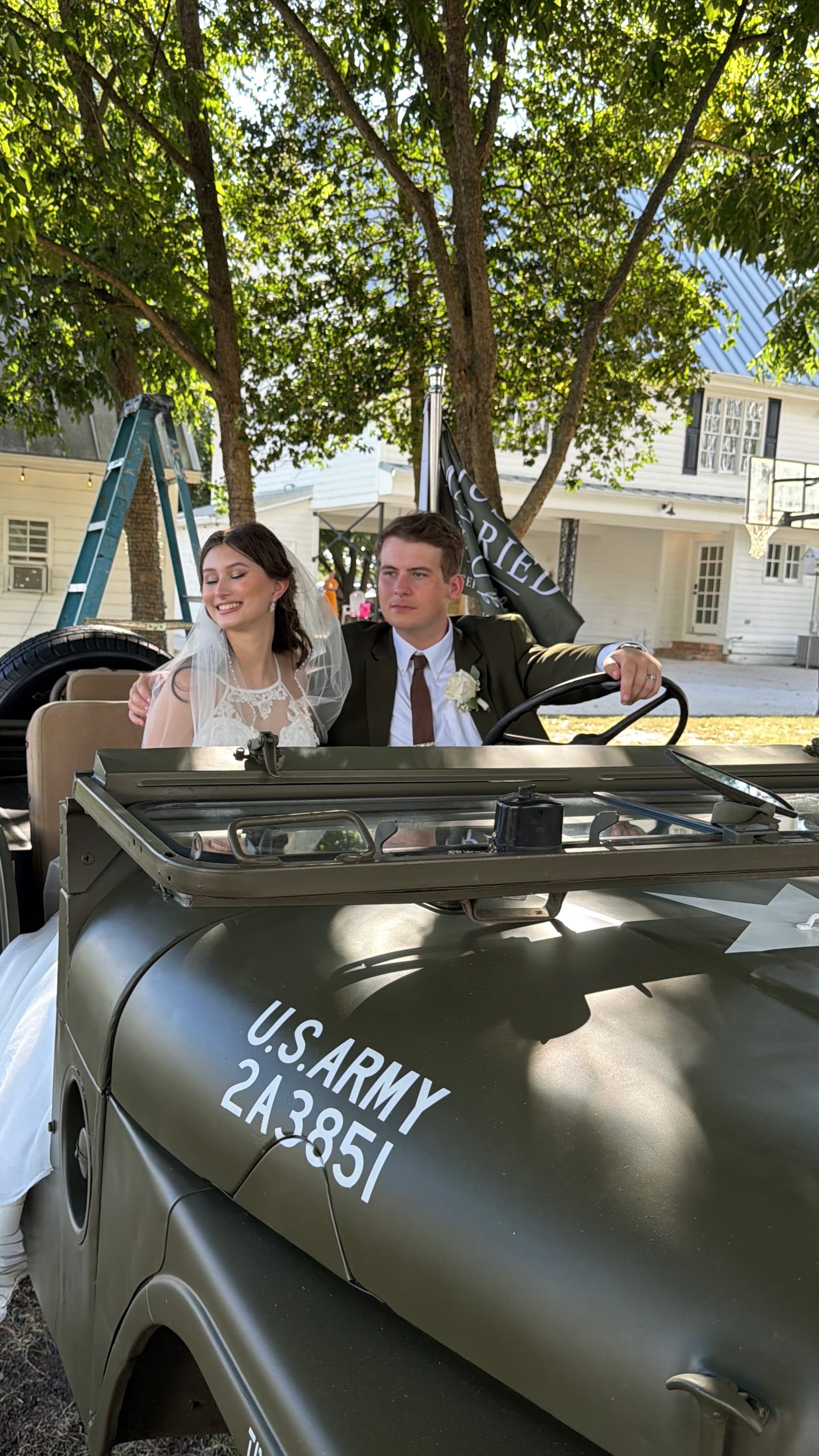 Bride in veil and groom in dark suit sit together in a vintage U.S. Army jeep in front of a historic white manor house after their wedding ceremony.