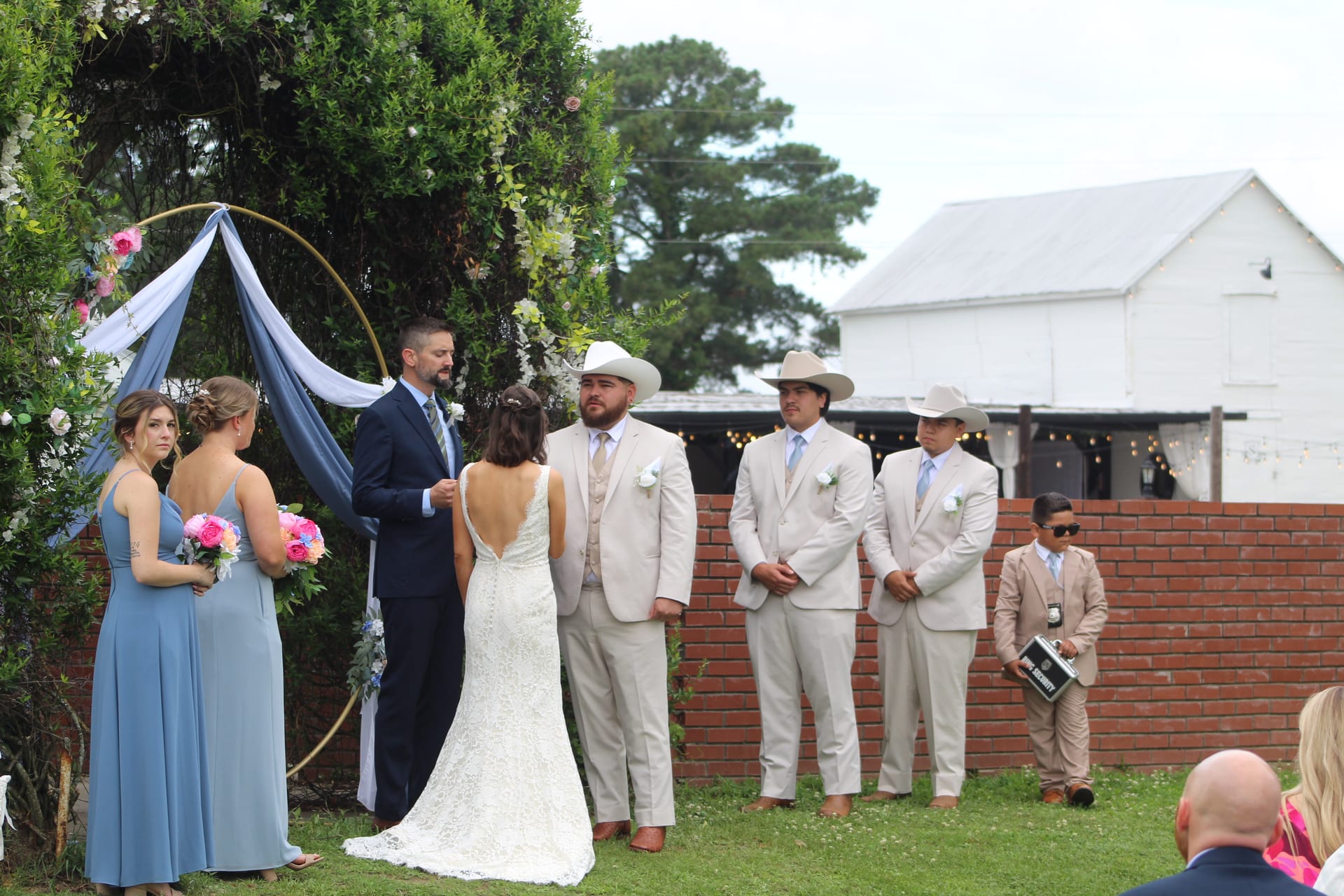 Bride and groom exchange vows beneath a floral hoop arch draped in dusty blue fabric on the ceremony lawn at Scarborough House, flanked by bridesmaids in blue gowns and groomsmen in tan suits with cowboy hats, with a white barn visible in the background.