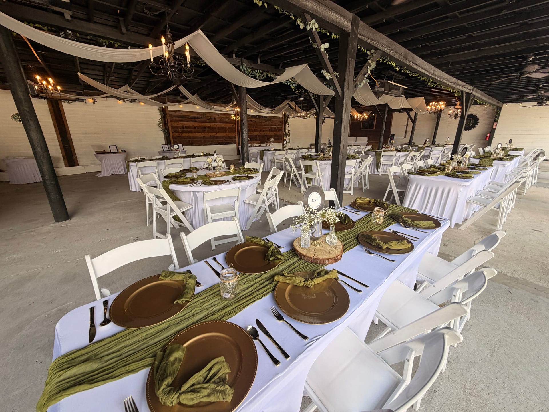 Elegantly set reception tables with white linens, gold charger plates, olive green napkins, and rustic wood centerpieces beneath a draped chandelier pavilion at Scarborough House.