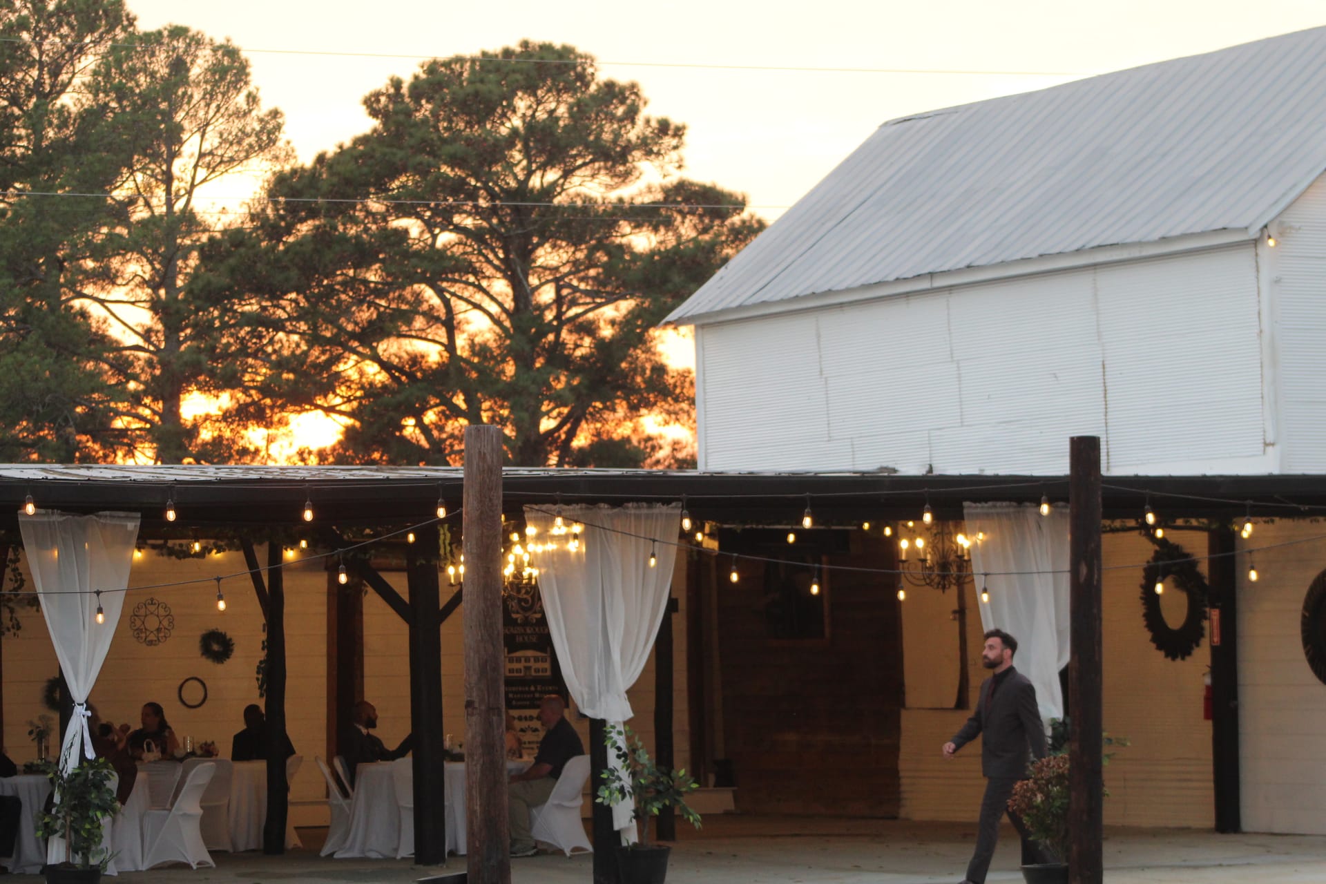 Guests gathered under a warmly lit rustic open-air pavilion adorned with string lights and white draping curtains at a historic barn wedding venue during a glowing golden-hour sunset.